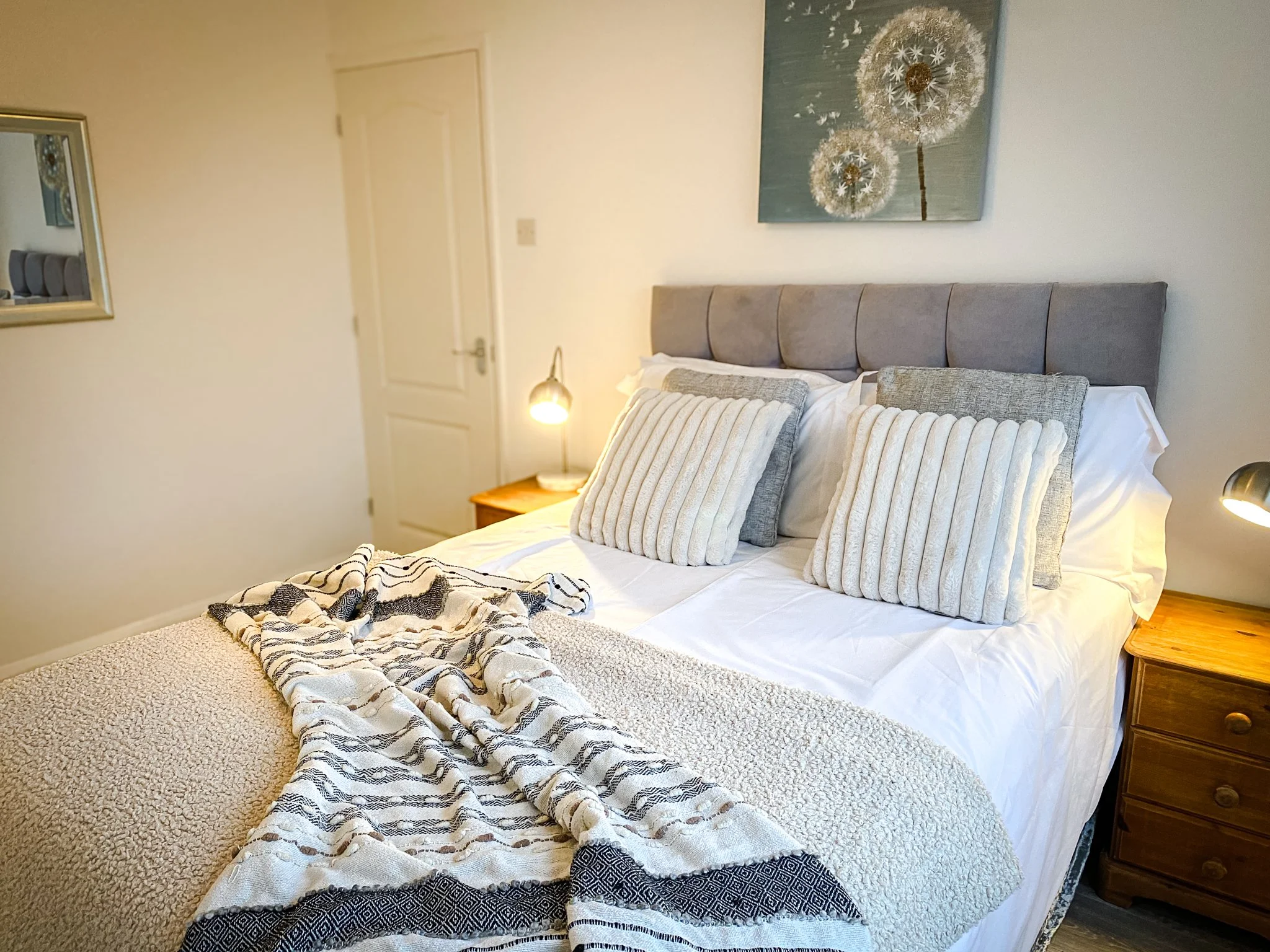 A neatly made bed with white sheets, beige and gray pillows, and a soft blanket, in a bedroom with a wooden nightstand, a small lamp, a mirror on the wall, and a painting of dandelions above the bed.