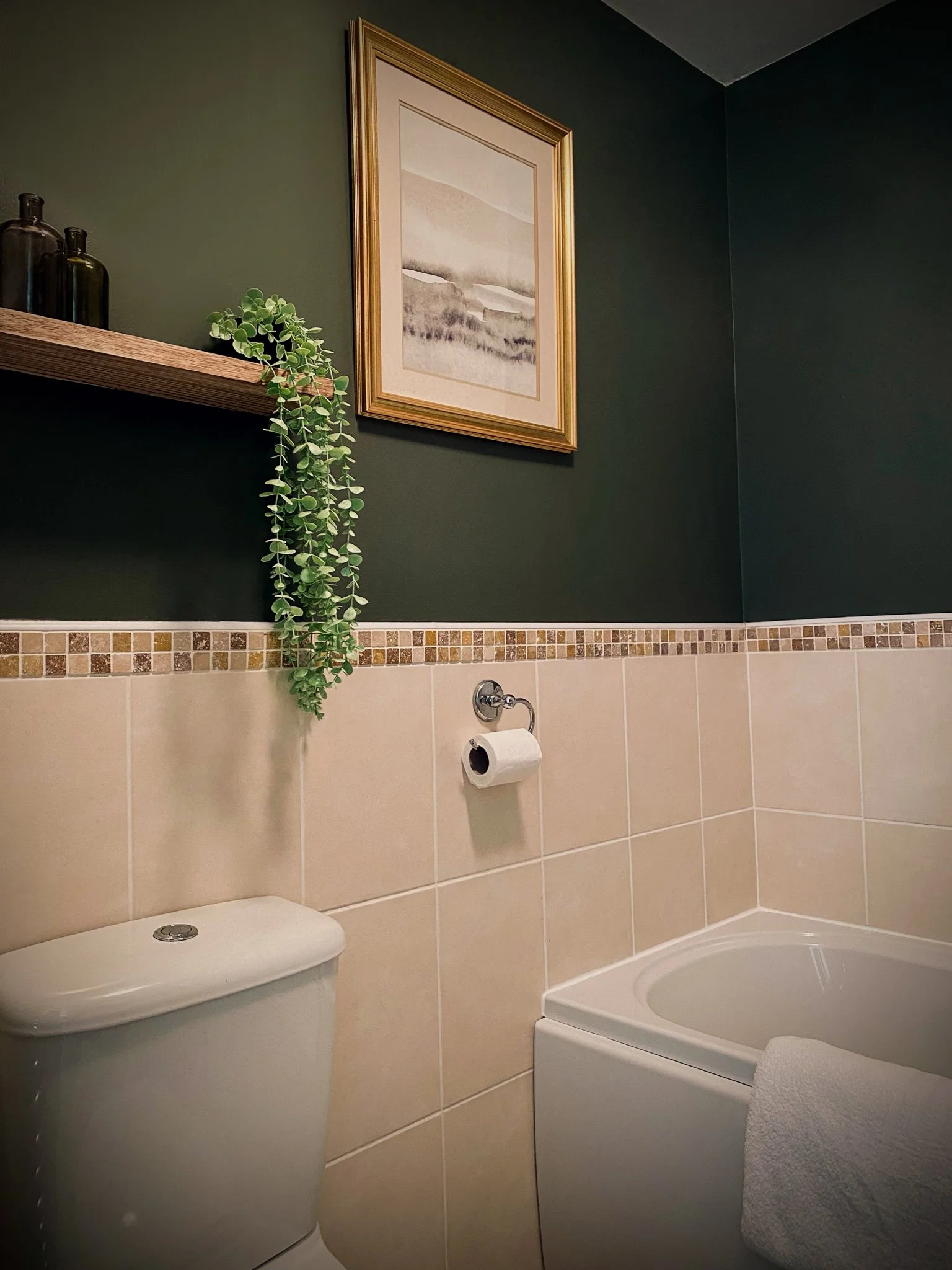 A bathroom with a toilet, a bathtub with a towel, a green wall with a framed landscape picture, a floating wooden shelf with decorative bottles and a hanging green plant, and a tiled wall.