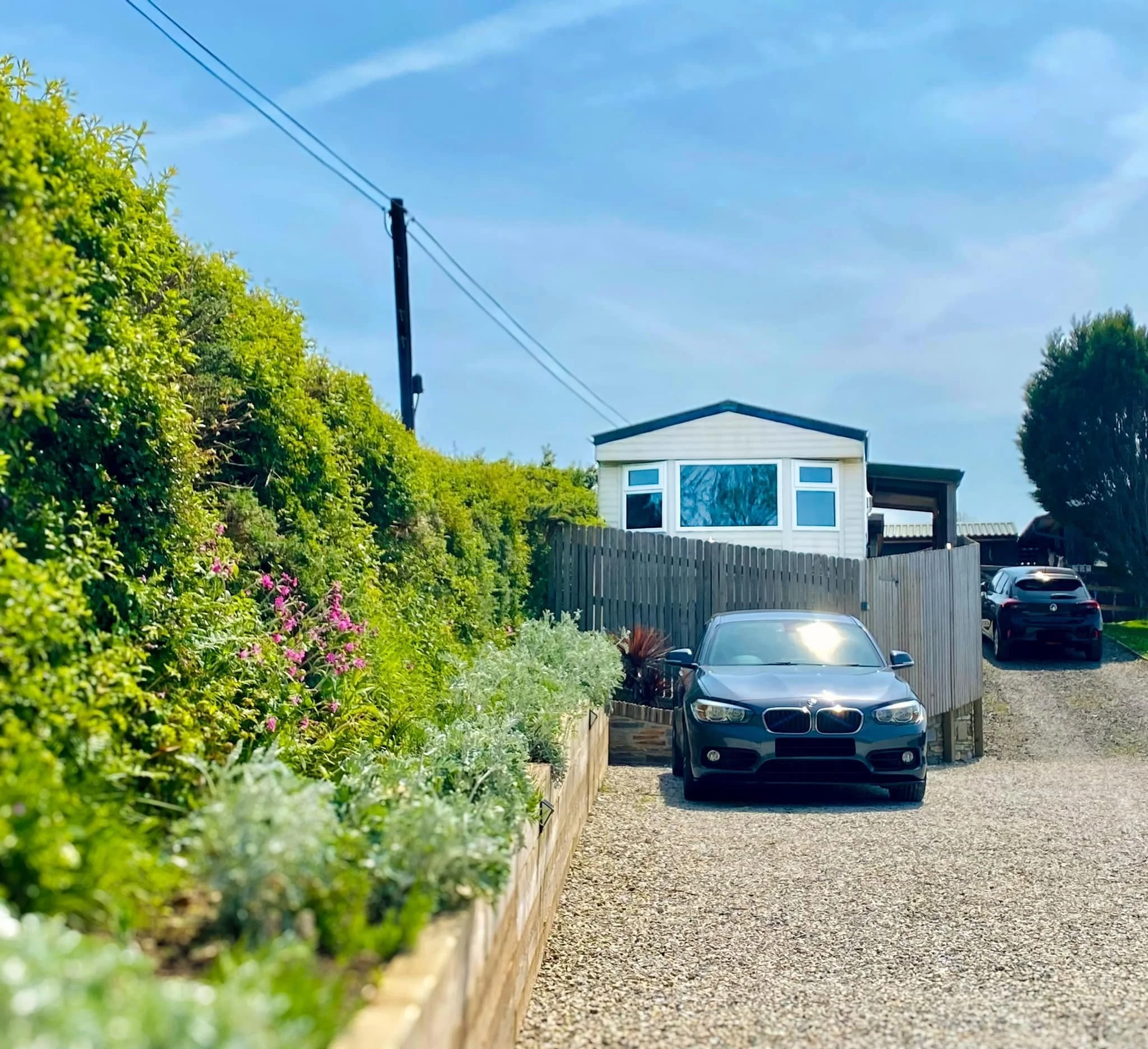 A gravel driveway with a black BMW parked near a wooden fence, leading to a house with a white exterior and large windows, surrounded by greenery and a clear blue sky.