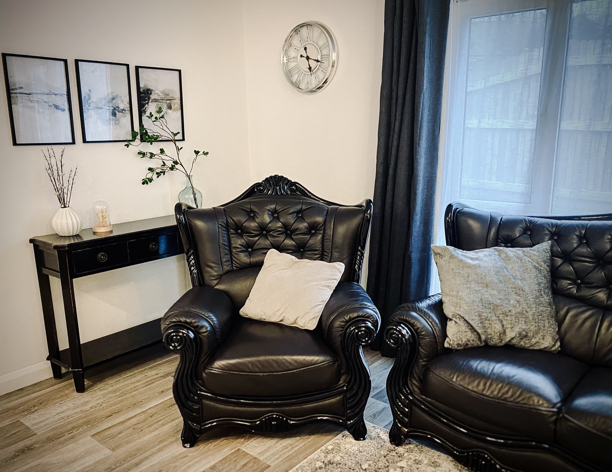 Living room with black leather armchair and sofa, pillows, wall clock, framed art, and side table with vases and decor, near window with curtains.