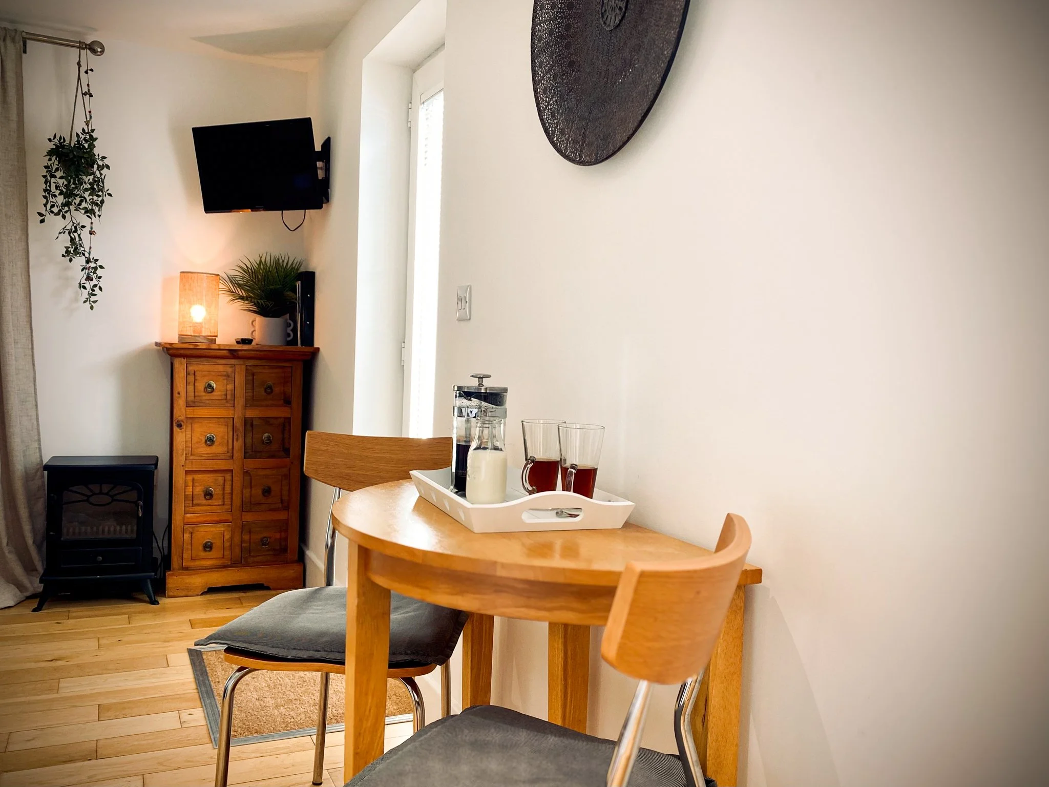 A small dining area with a wooden table and two chairs, set with a tray holding two glasses of tea, a French press, and a bottle of milk. In the background, a wooden cabinet with a lamp, a potted plant, and a small black appliance beneath a televisio