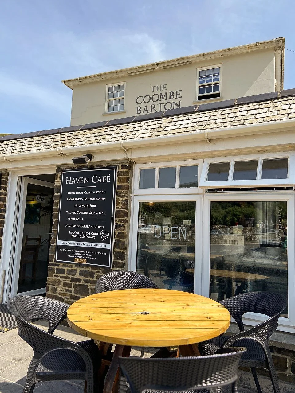Exterior view of Haven Café with a blackboard menu and outdoor seating with a round wooden table and black chairs, a building with a sign for The Coombe Barton on the upper floor, and a blue sky.