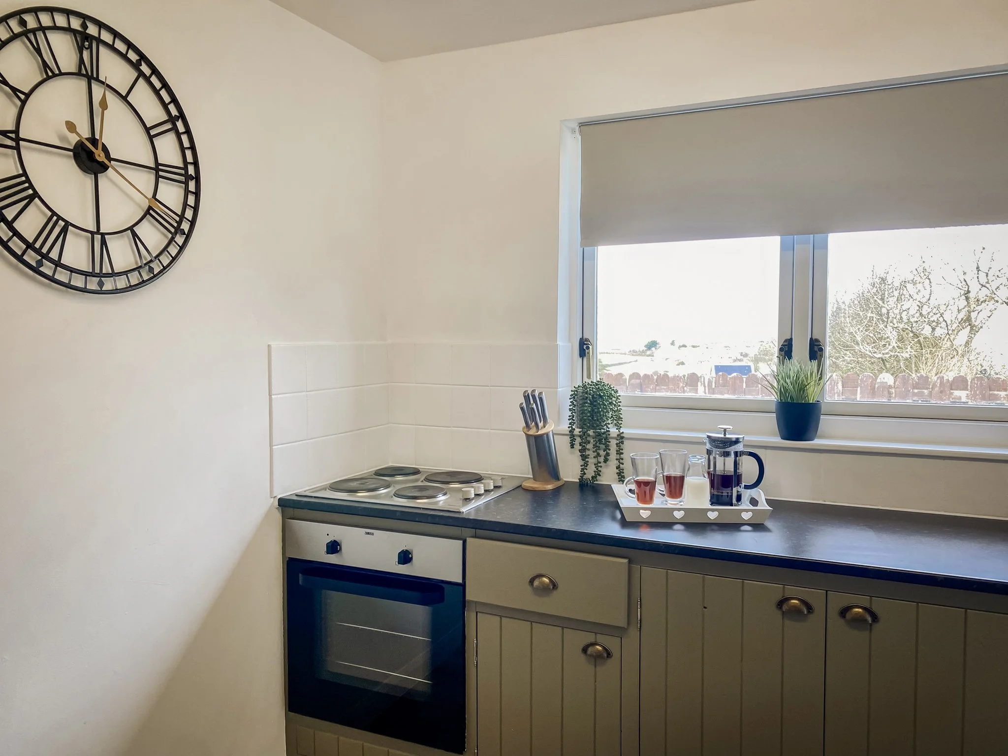 Kitchen corner with a window, clock on the wall, stove, countertop with potted plants, glassware, and a French press.