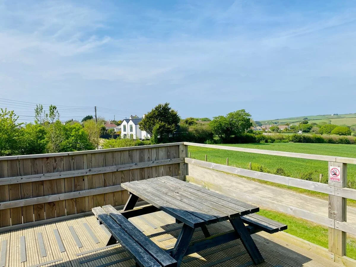 Wooden deck with picnic table and railing overlooking a grassy countryside with houses and trees under a blue sky.