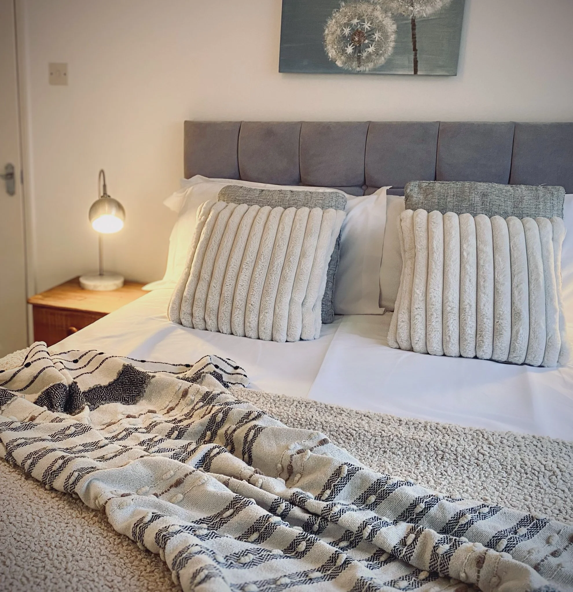 A neatly made bed with beige and gray pillows, a cozy blanket, and a gray upholstered headboard in a bedroom. A small bedside table with a lamp is visible on the left, and a decorative artwork hangs above the bed.