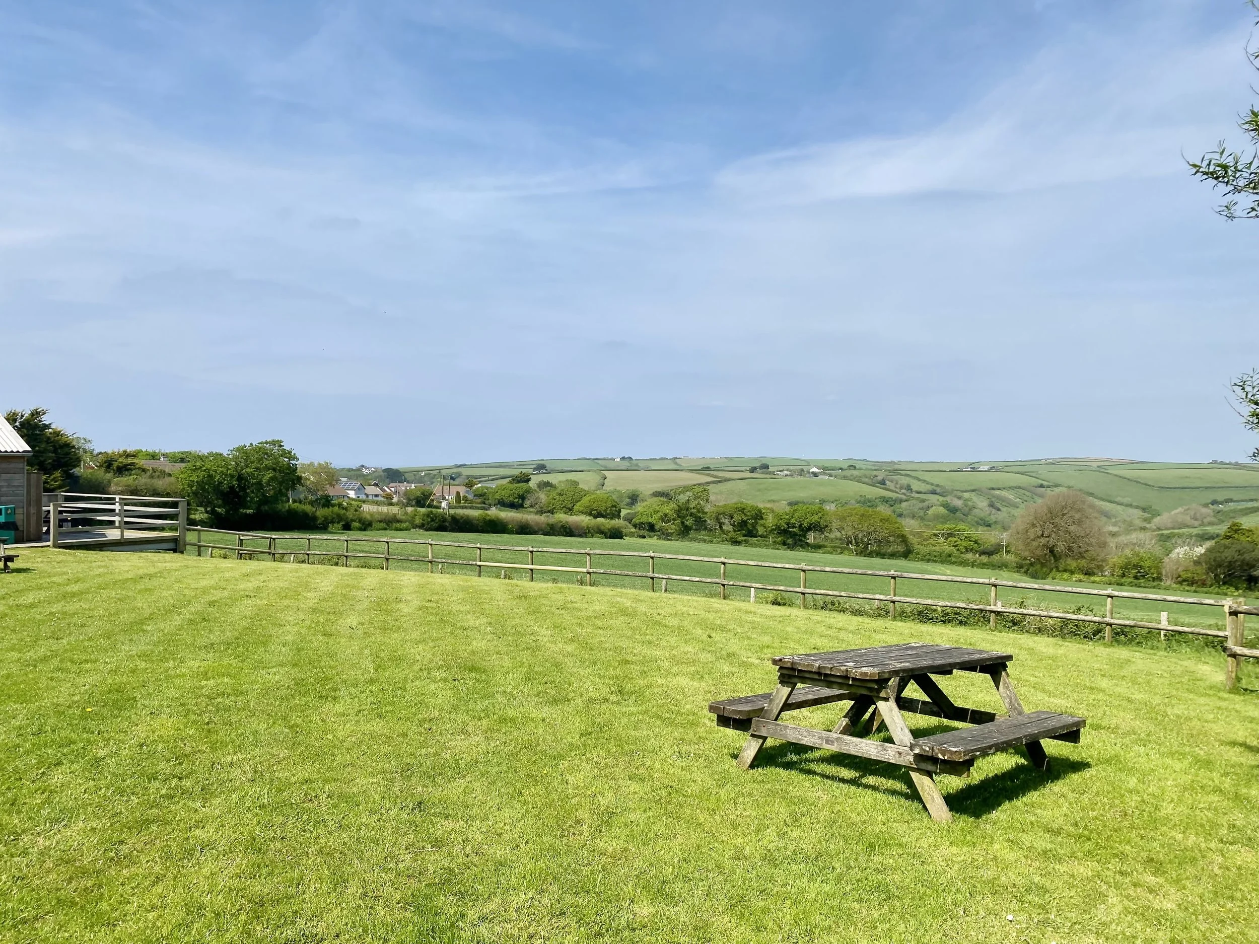A grassy field with a wooden picnic table, a fence, and rolling green hills in the background under a partly cloudy sky.