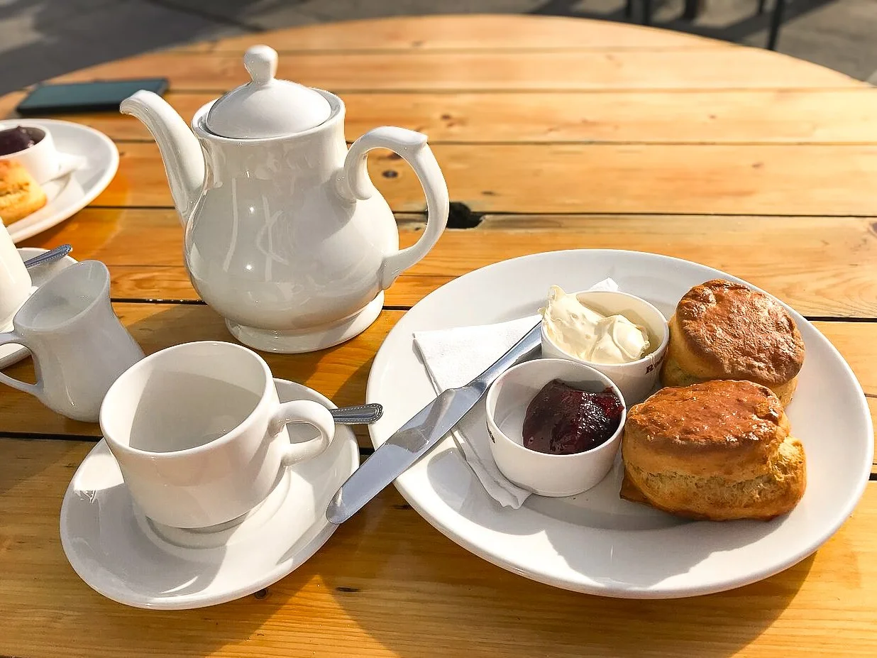 Tea set with a teapot, teacup, and creamer on a wooden table. A white plate with two scones, clotted cream, and jam.