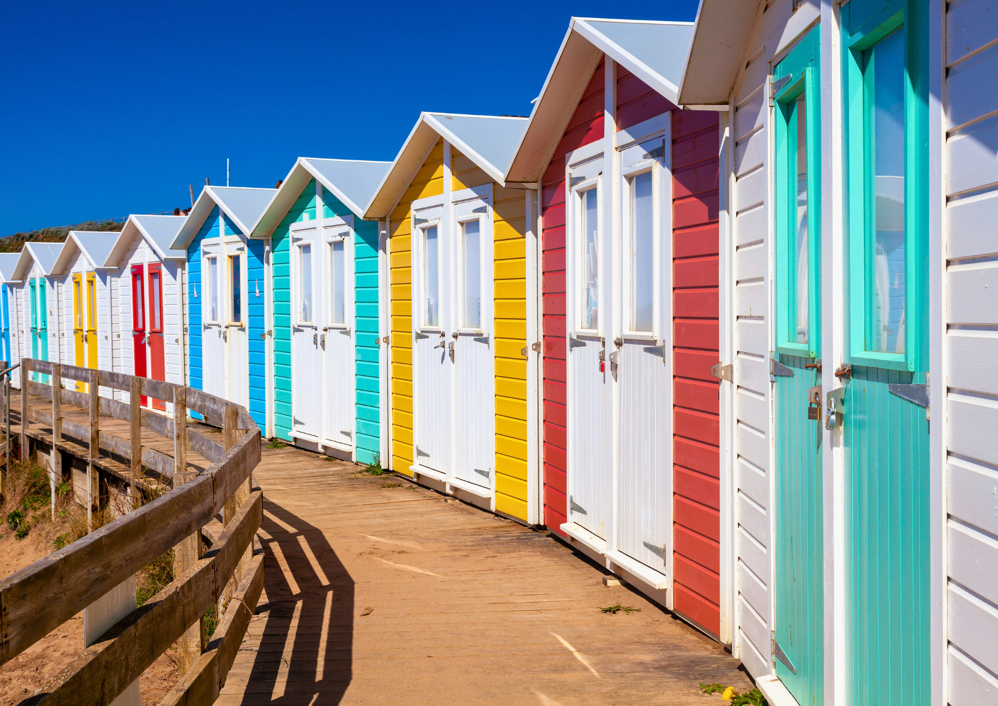 Colorful beach huts lined up along a wooden walkway on a sunny day with a clear blue sky.