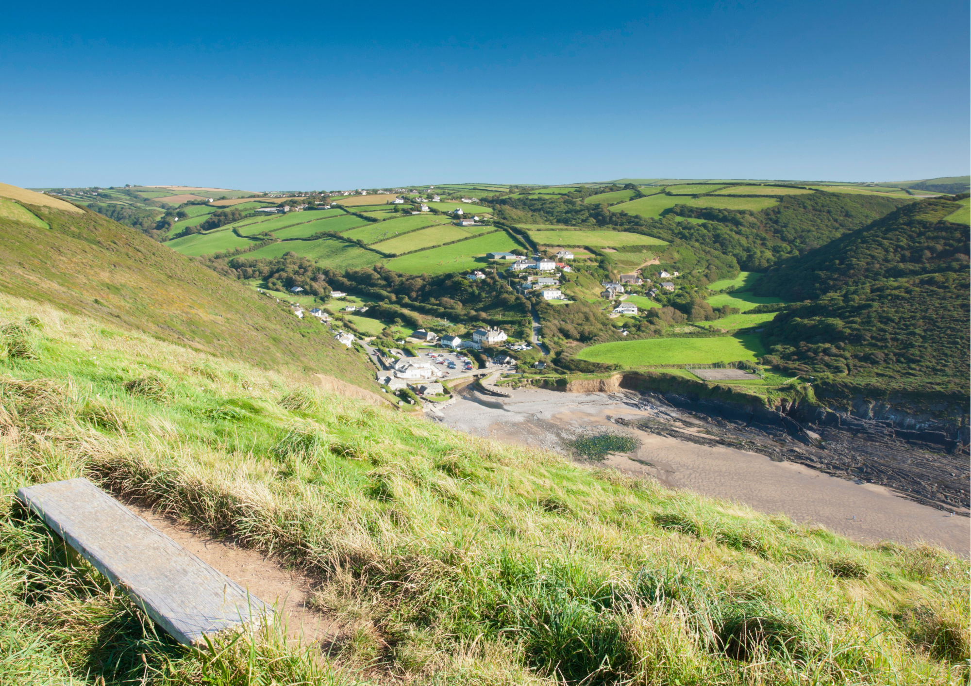 A vibrant landscape of rolling green hills and a small coastal village with white houses and narrow roads, overlooking a sandy beach and the ocean under a clear blue sky.
