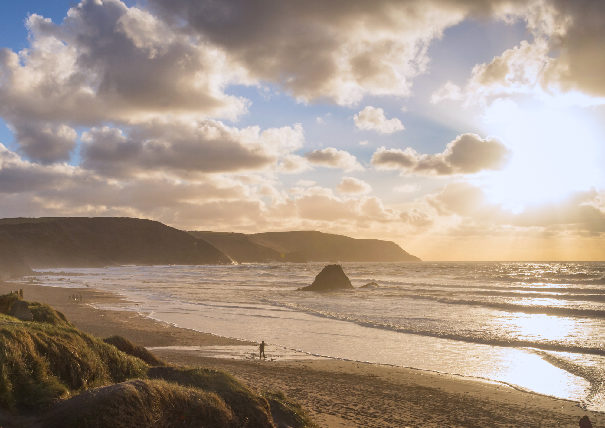 Sunset over a beach with clouds, ocean waves, and rocky cliffs in the distance.