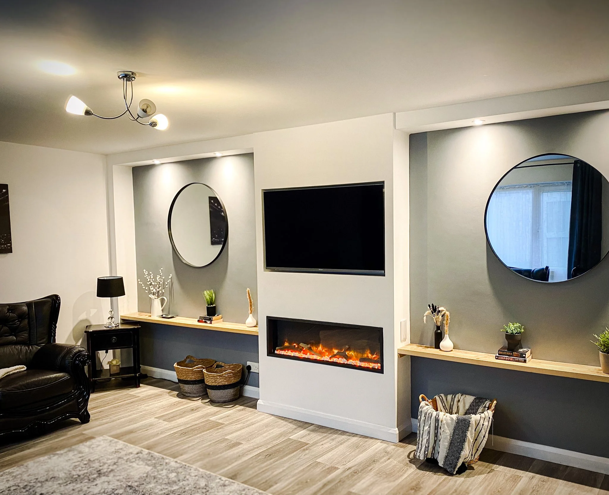 Living room with a mounted flat-screen TV, an electric fireplace, circular mirrors, a leather armchair, decorative plants, and baskets, with modern lighting.