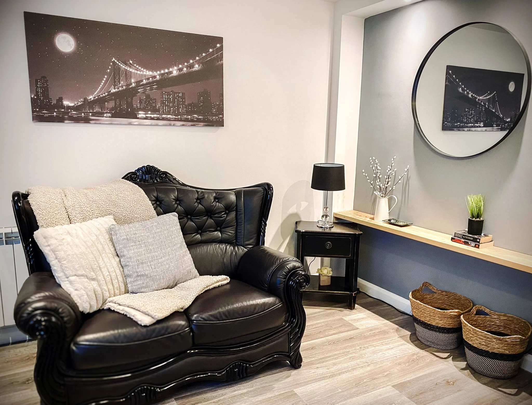 Living room with black leather tufted sofa, pillows, a side table with a black lamp, wall art depicting a bridge at night, and woven baskets on the floor.