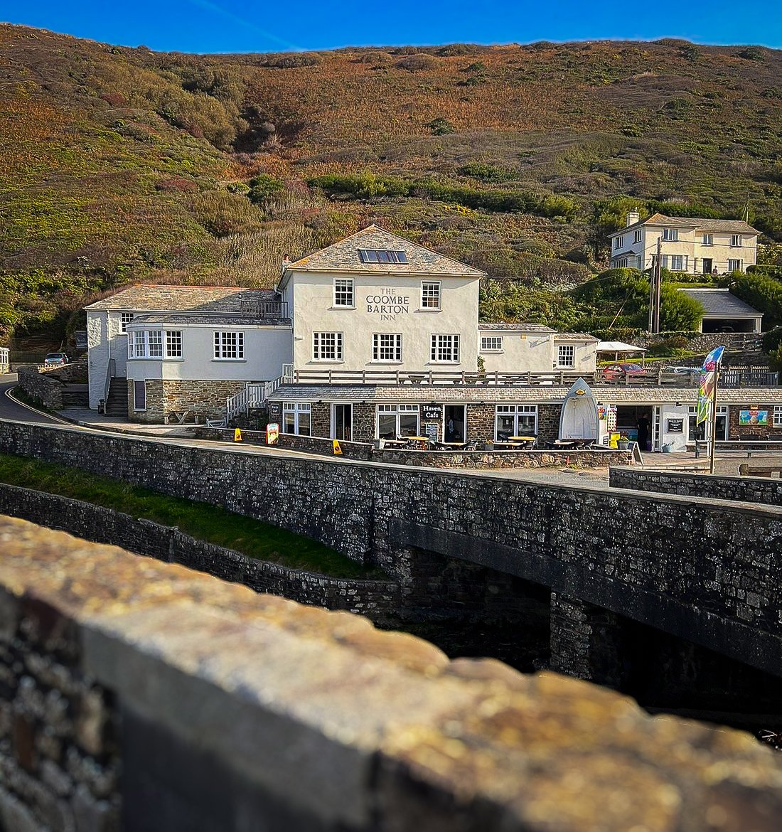 A white building with multiple windows, labeled 'The Coombe Barton Inn,' situated behind a stone wall with a seating area outside, set against a hillside with greenery and other houses.