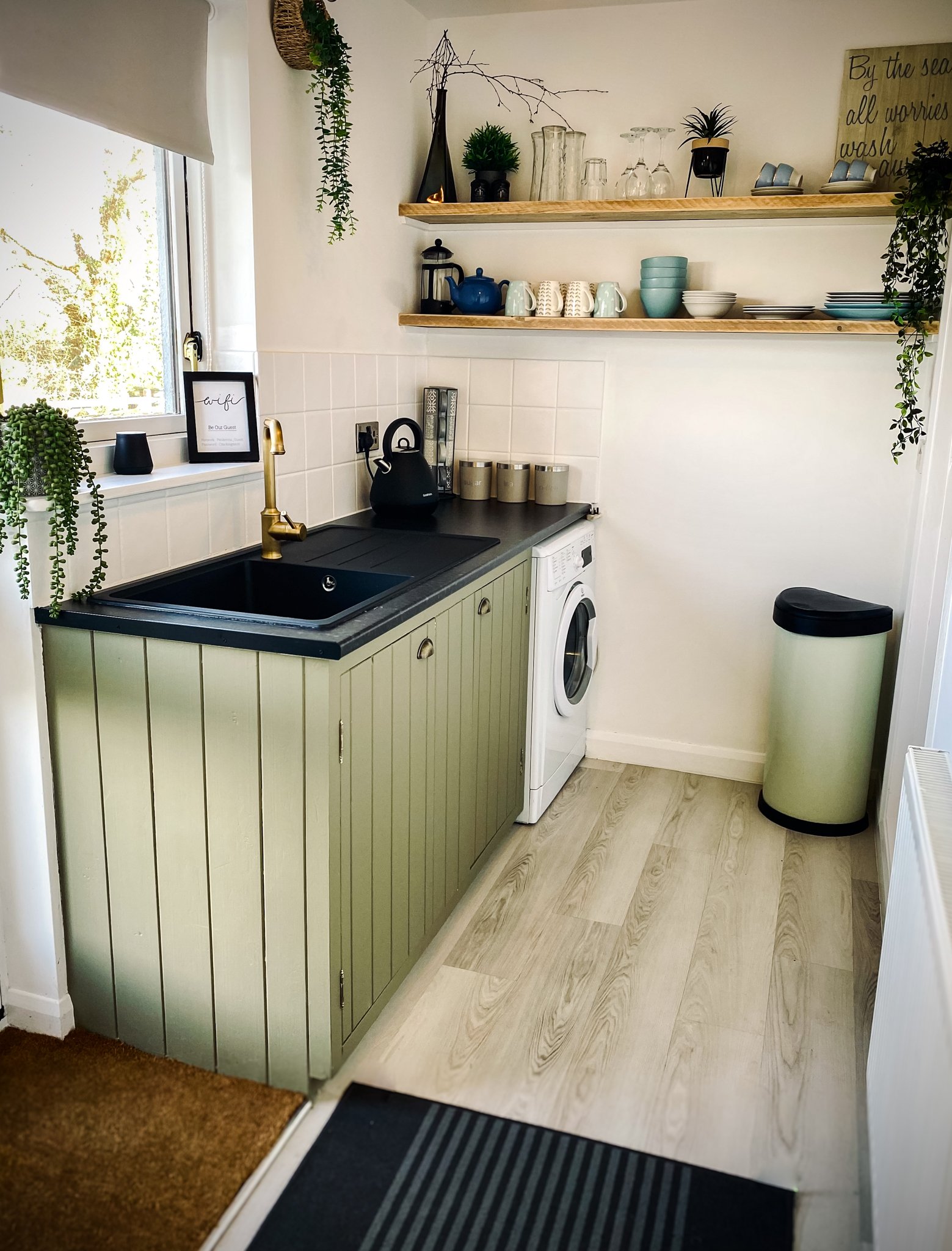 A small kitchen corner with a black sink, a gold faucet, a washing machine, and two wooden shelves with dishes, cups, and decorative items. There is a window with a white blind, and hanging plants. A black kettle and beige canisters are on the counte