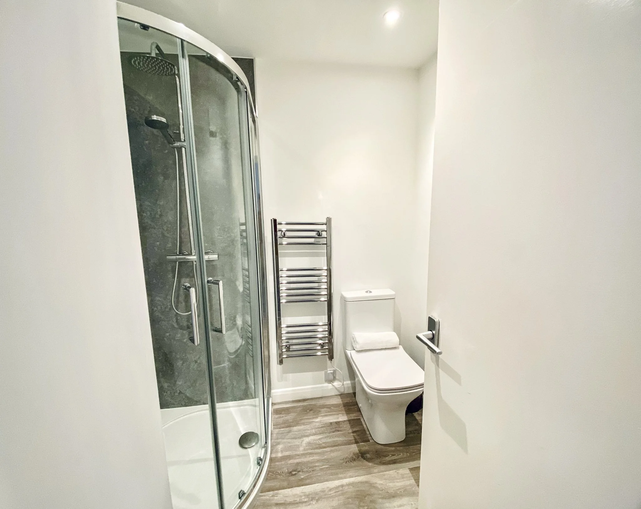 Small modern bathroom with a shower enclosure, a toilet with a towel on the lid, a heated towel rail, and light-colored walls and floor.