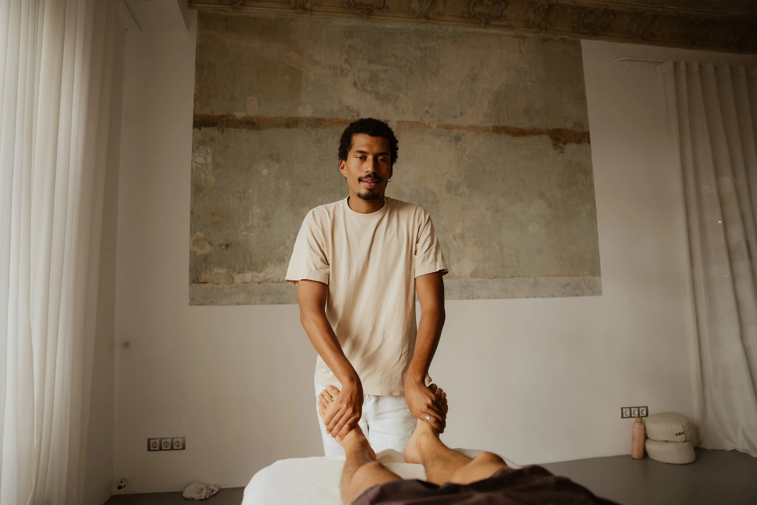 A man receives a massage from a massage therapist in a room with a beige wall and minimal decor.
