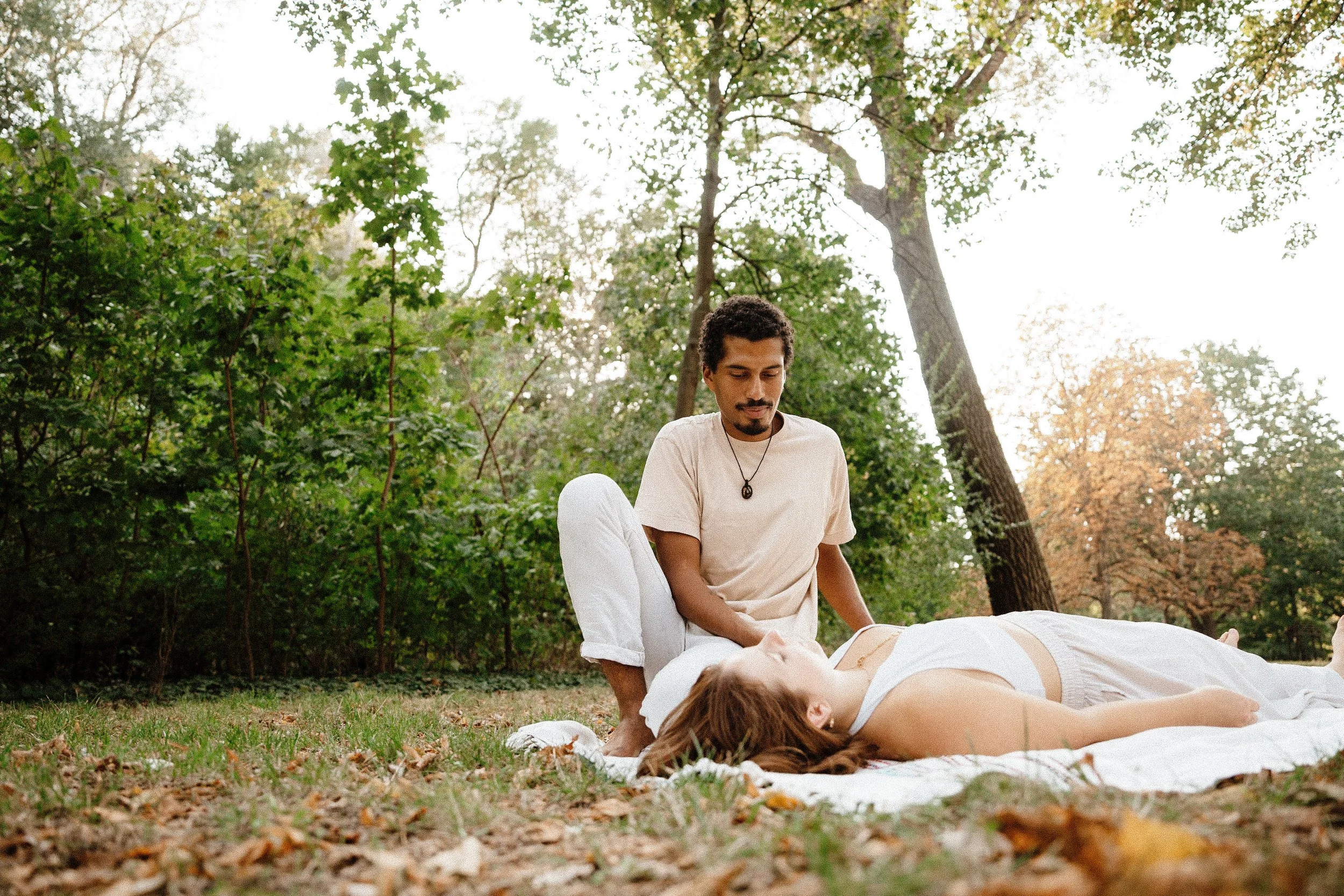 A man practicing integrative Bodywork healing on a woman lying on a white blanket in a park surrounded by trees.