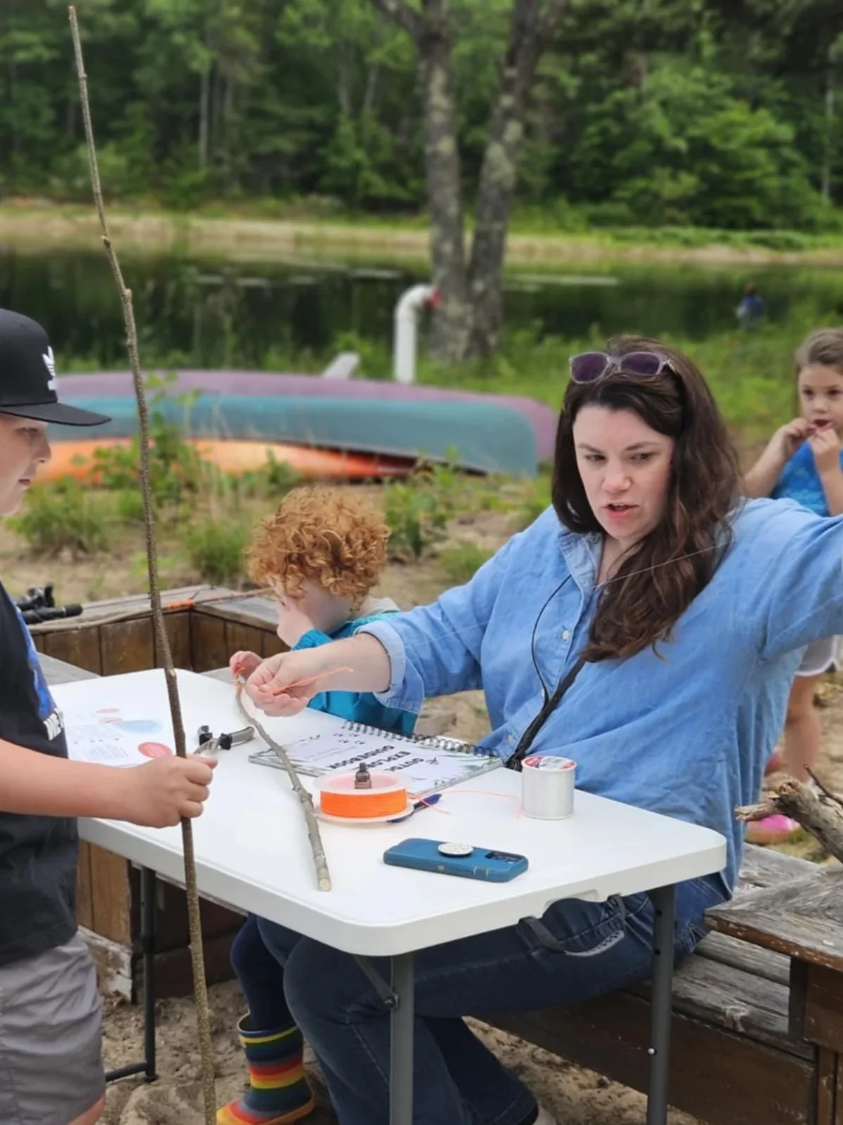 Speech Therapist is outside sitting next to a small child while explaining how to make your own fishing pole to an older child.