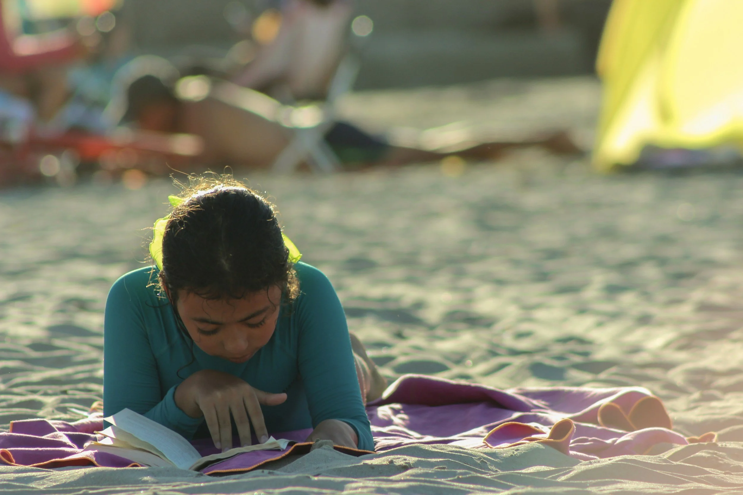 A person with dark hair in a blue shirt is lying on a beach towel on the sand, reading a book with sunlight shining behind.