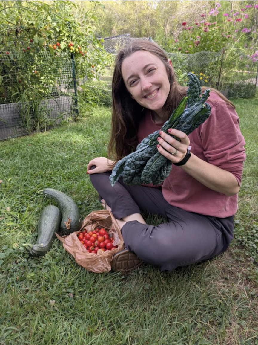 Julie, a speech clincian, is smiling and sitting on a grassy lawn, while holding a bundle of green leafy vegetables.