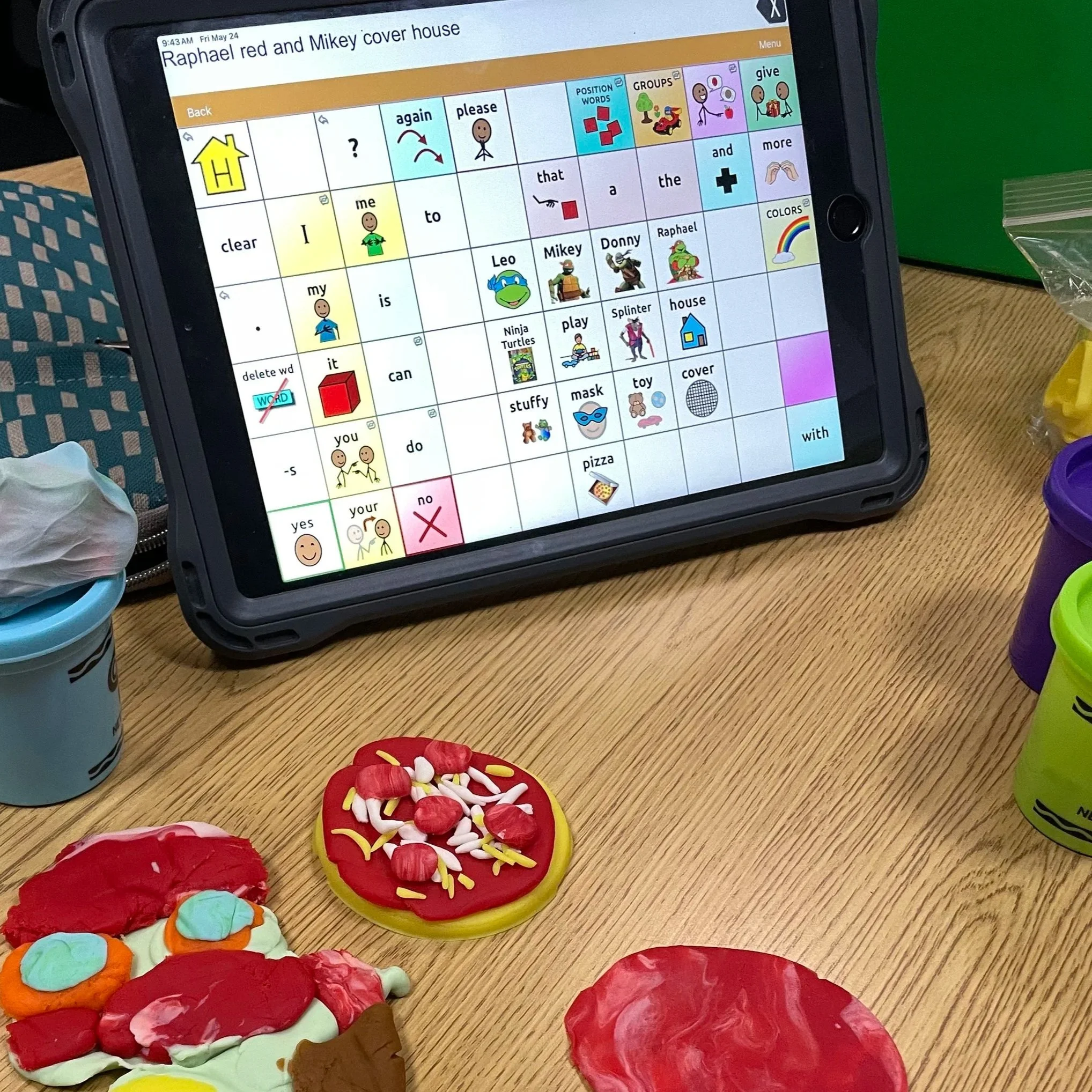 A tablet, showing a speech-generating AAC system, is sitting on a table with pretend pizza and playdough.