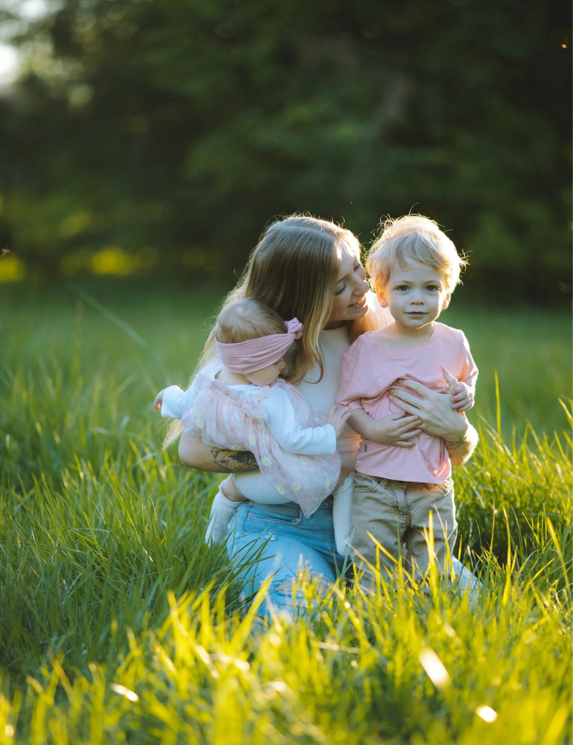 A woman with long blonde hair holding two young children in a grassy field during golden hour, surrounded by trees.