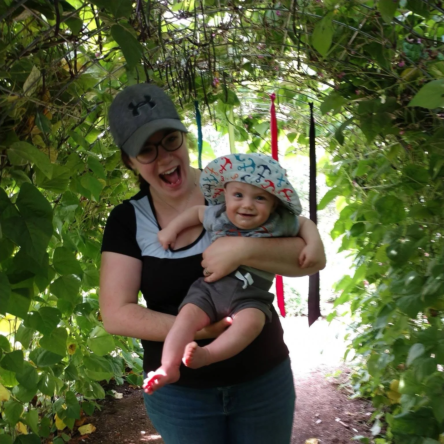 The Found of Chickadee Chatter, Katherine Rackliff, smiling while holding a small child in a brightly, lit tunnel of vines.