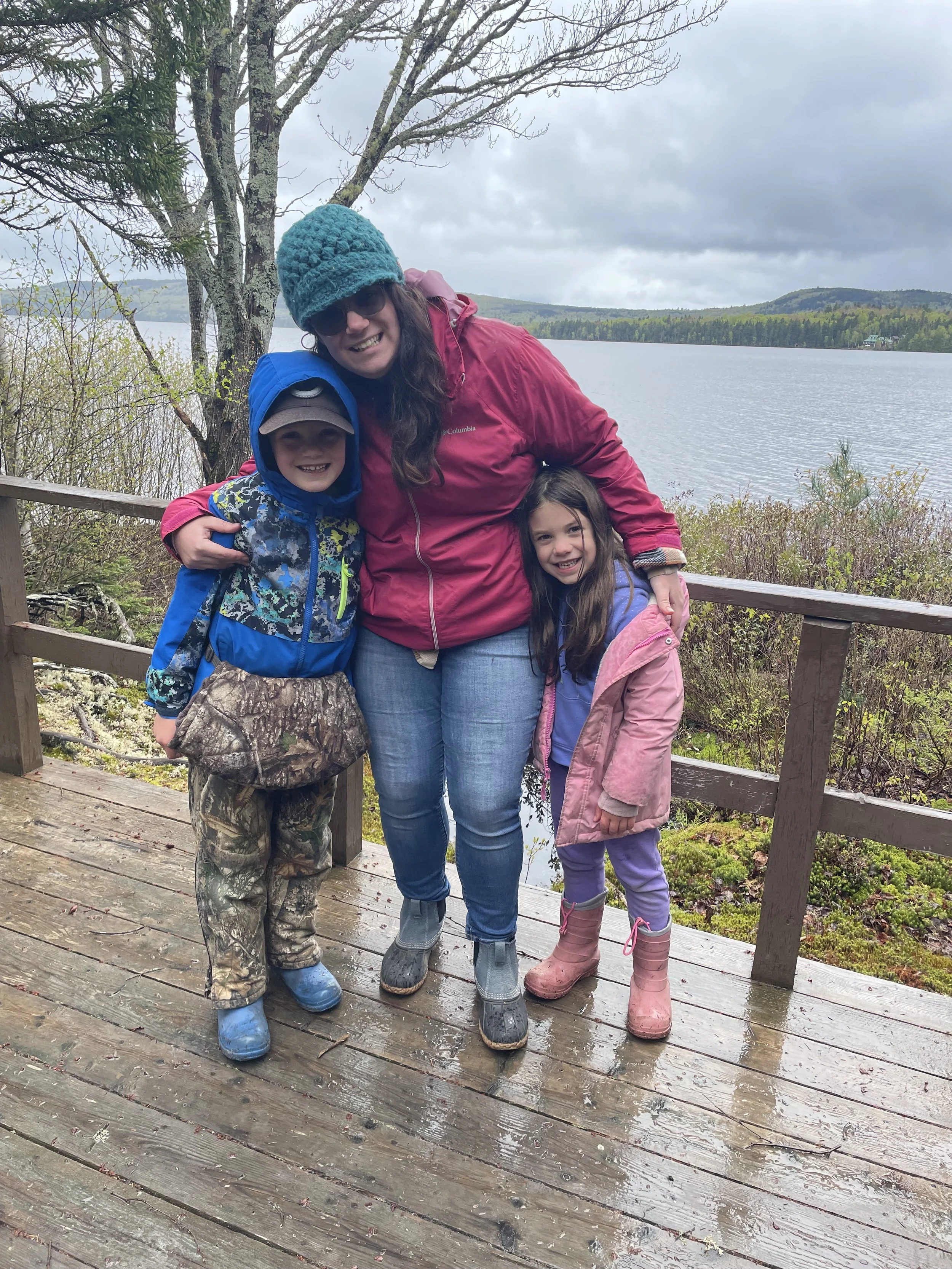 A woman and two children standing on a wooden dock near a lake, dressed in rain jackets and boots. The woman is wearing a red jacket, a teal knit hat, sunglasses, and has long brown hair. The boy is wearing a blue raincoat, camouflage pants, and a brown cap. The girl is wearing a purple jacket, lavender pants, and pink rain boots. They are smiling and posing for the photo, with a lake, trees, cloudy sky, and hills in the background.