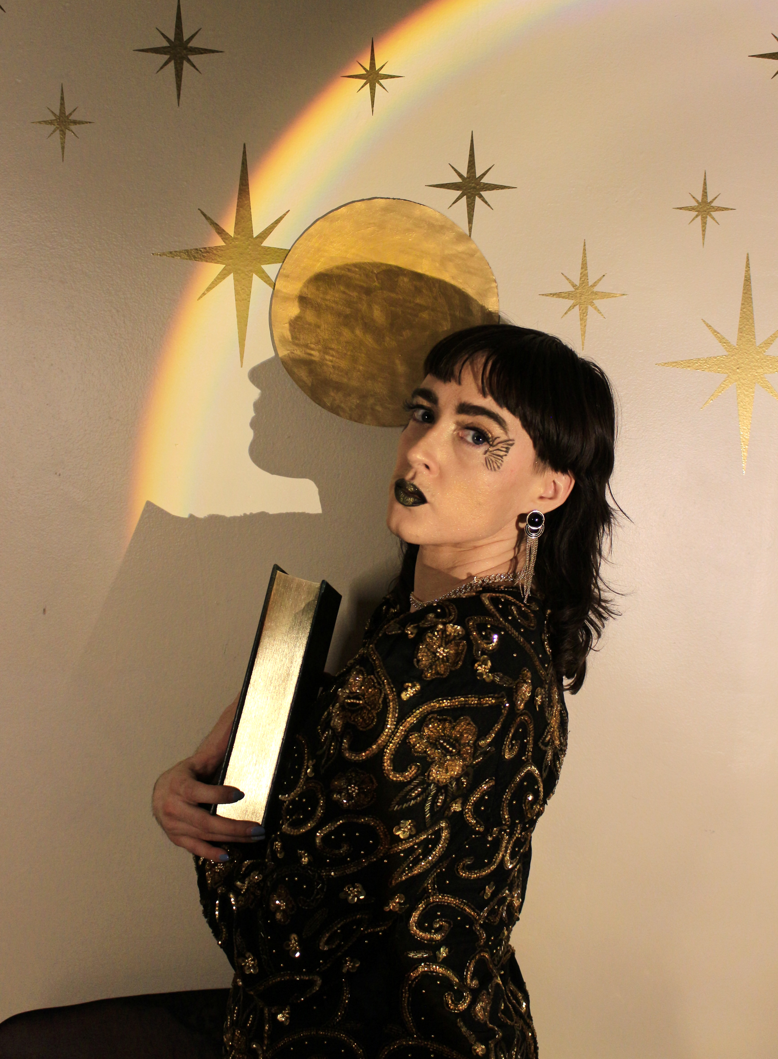 Aryn the Alchemist, a white person with a brown mullet, is wearing a gold and black shirt, black and gold earrings, and black makeup shaped like a butterfly near their face.