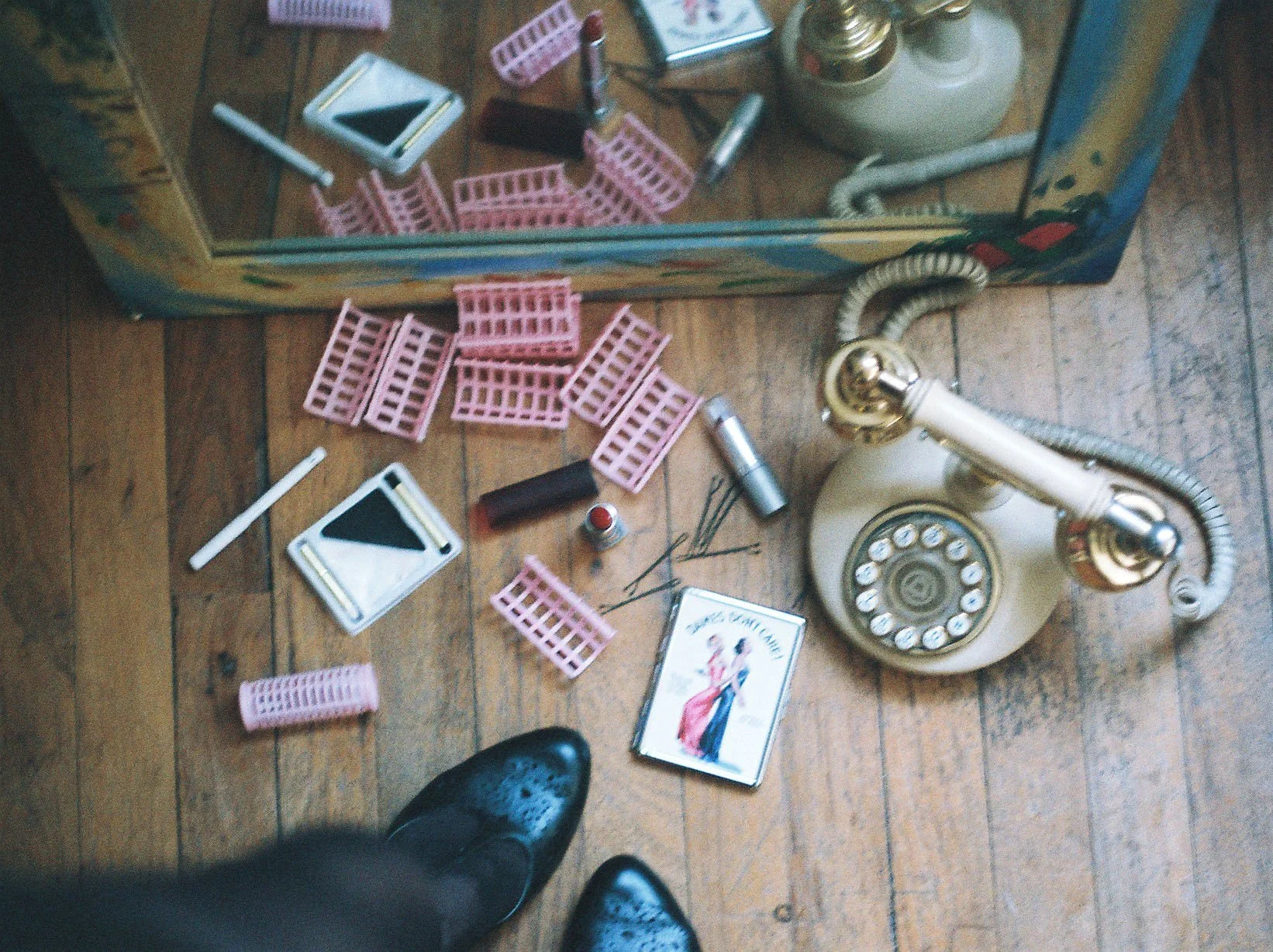A film image taken by Aryn Scanlon of black shoes, pink hair curlers, white and black makeup, a lipstick and a vintage telephone on a wooden floor.