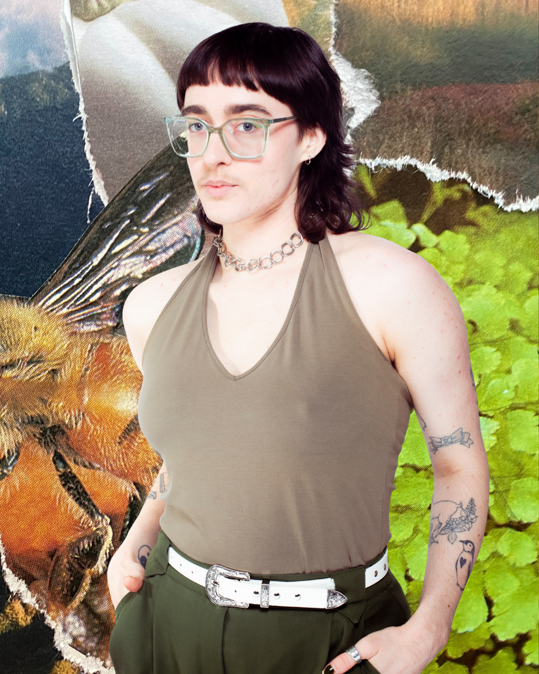 Portrait of Aryn the Alchemist, a white person with green glasses and a brown mullet wearing a green halter top. They are posed in front of an original collage.