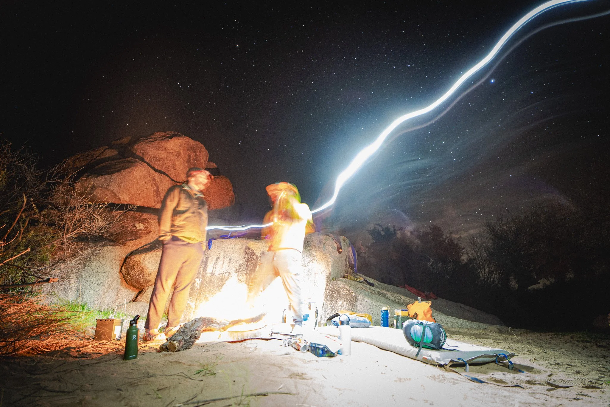 Two people standing on rocky ground at night with starry sky and lightning streak in the background; camping gear scattered around.