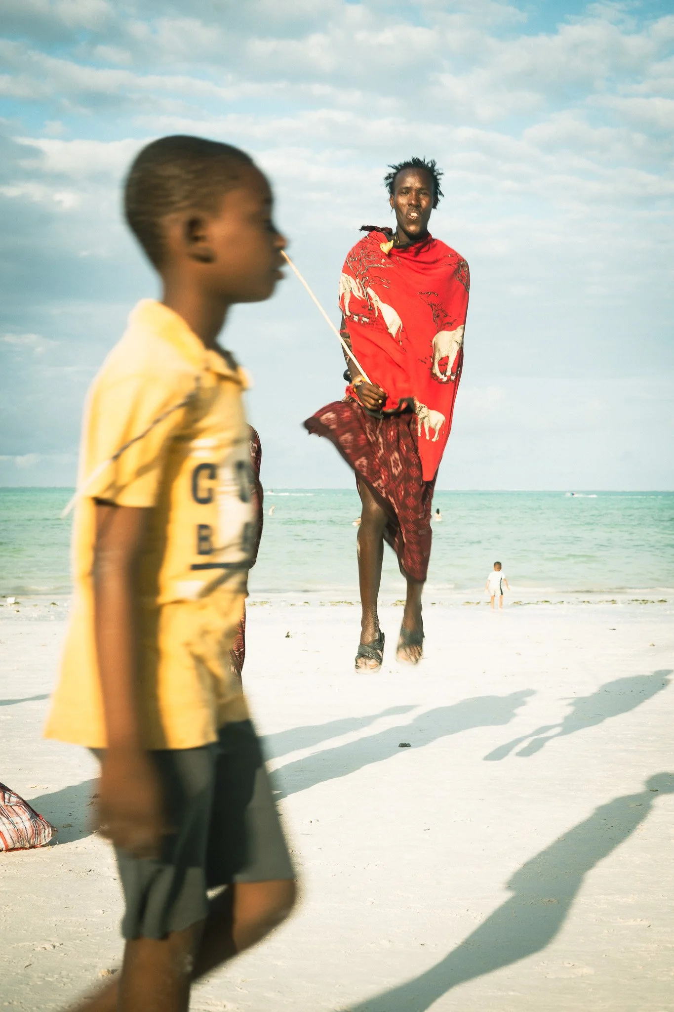 Two boys on a beach with a man jumping rope in the background, the ocean and sky visible behind them.