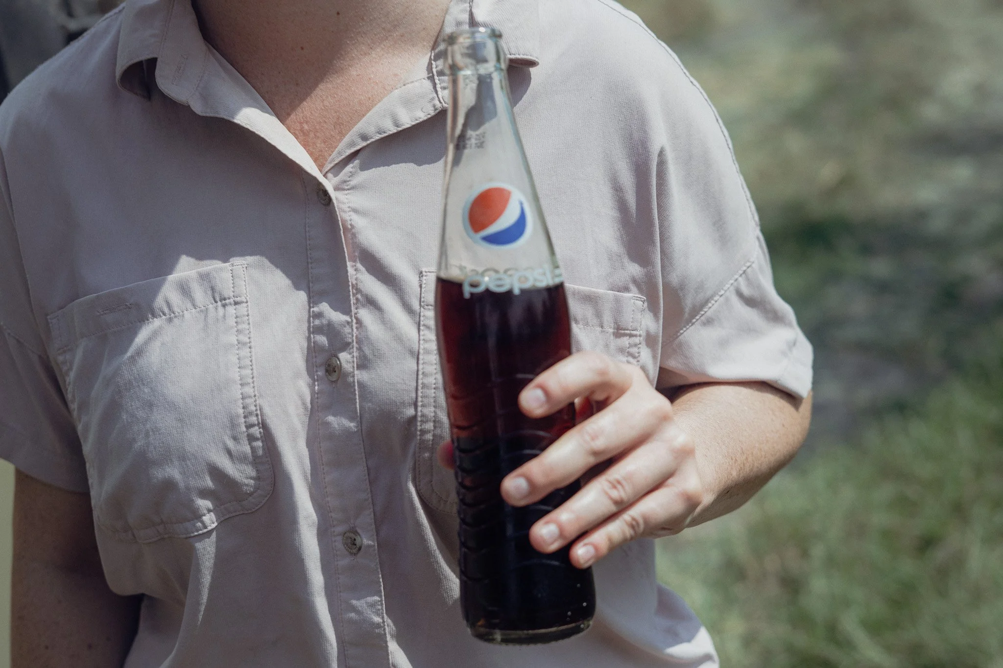 Person holding a glass bottle of Pepsi outdoors, wearing a light-colored short-sleeve shirt.