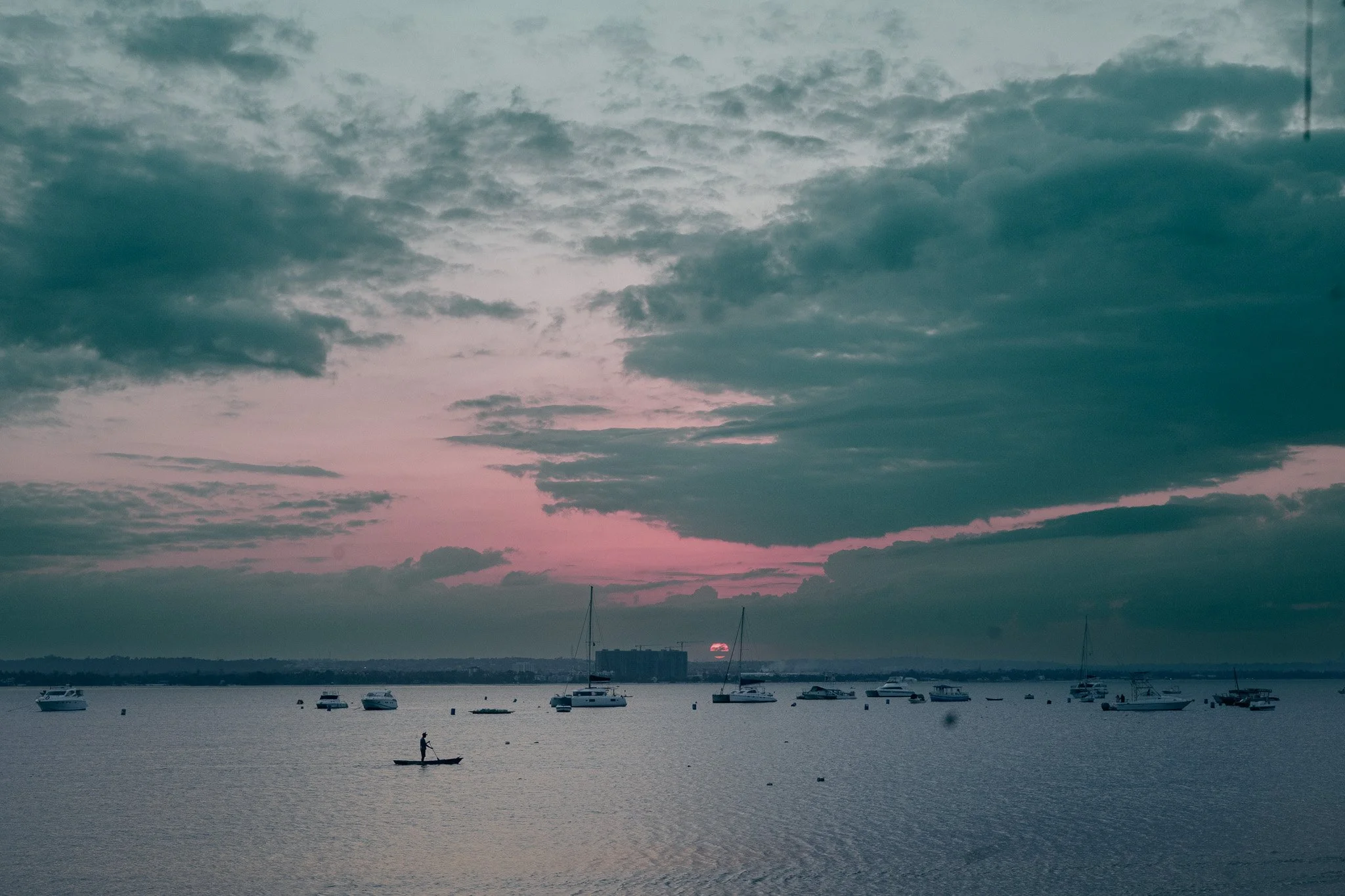 Sunset over a calm body of water with boats and a person paddleboarding, cloudy sky with pink and blue hues.
