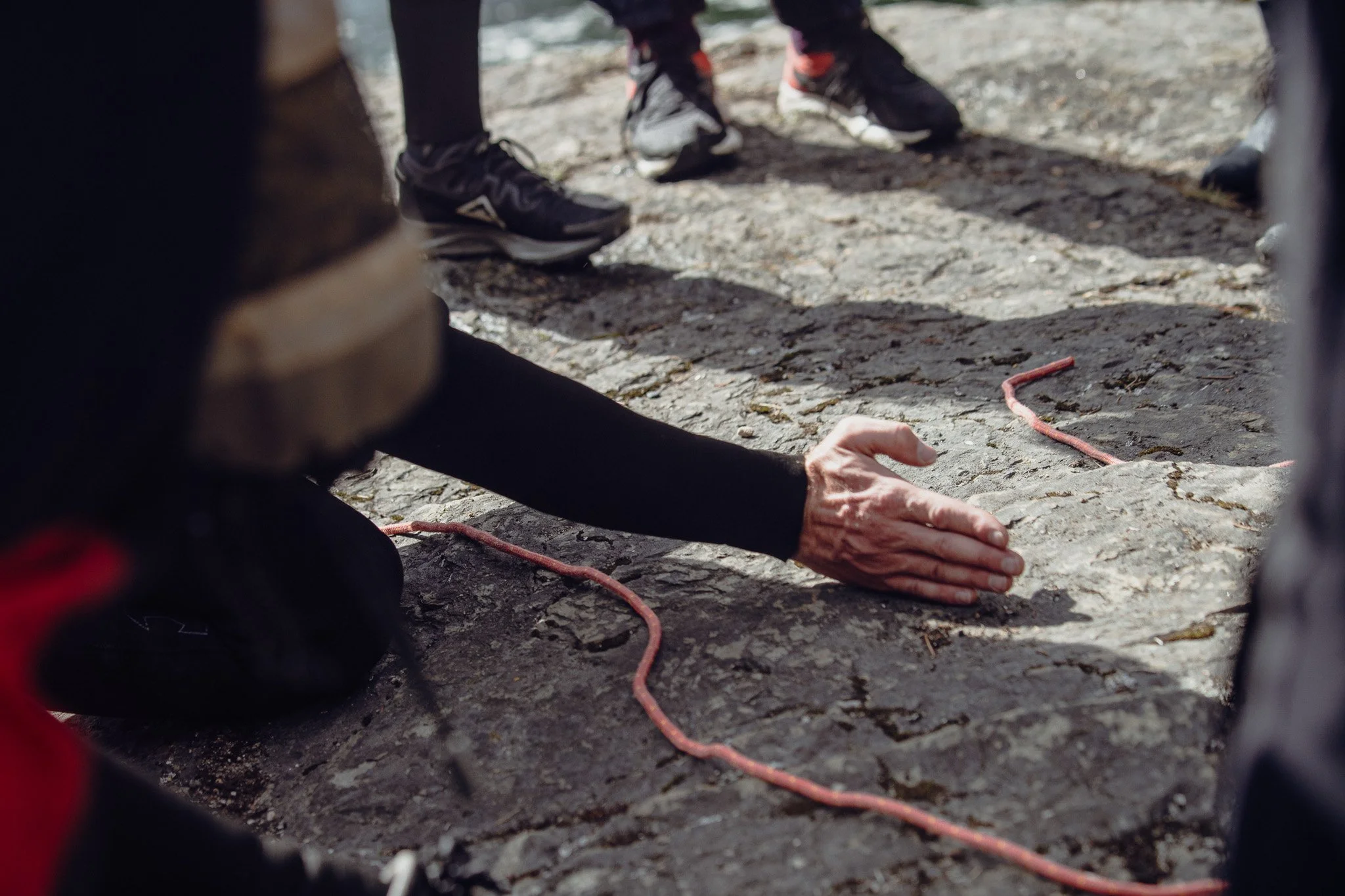 A person lying on the ground with their hand extended, participating in a tactile or sensory activity on a rocky surface, with other people standing nearby.