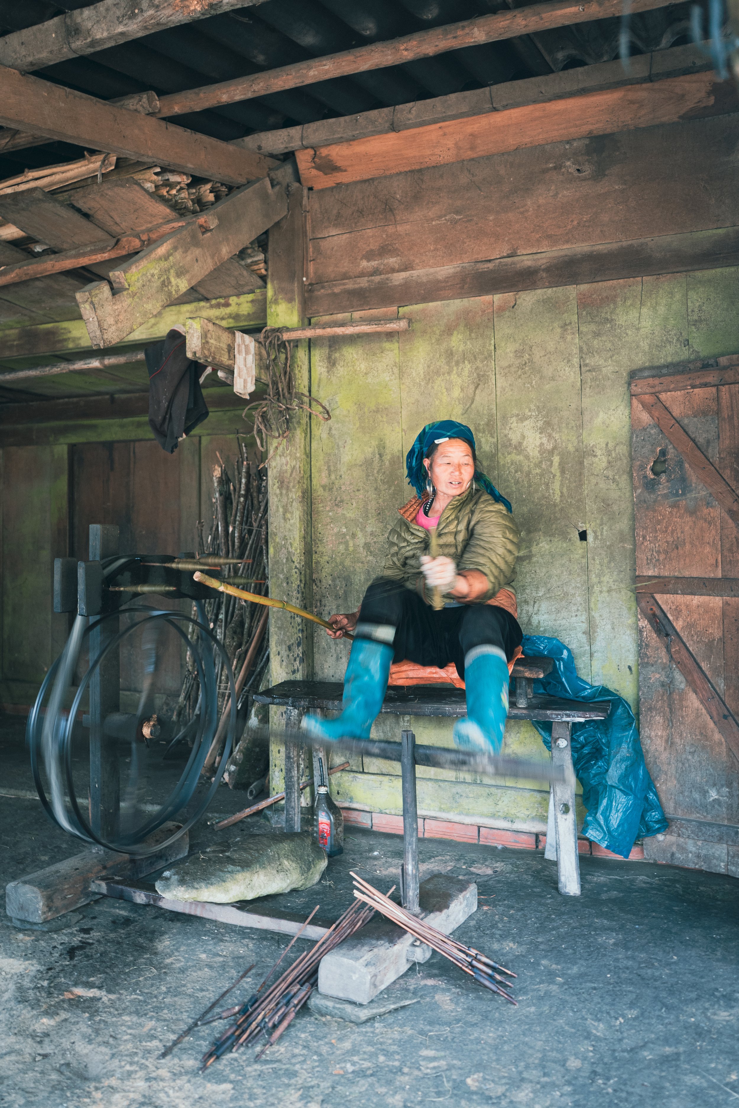 An elderly woman sitting on a bench inside a rustic wooden shed, wearing blue rain boots and holding a long stick, with various tools and objects around her.