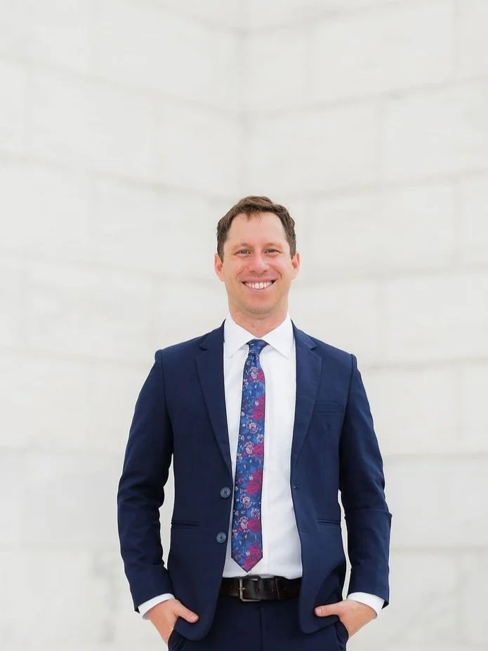 Smiling man in a blue suit, white shirt, and floral tie, standing in front of a plain light-colored wall.
