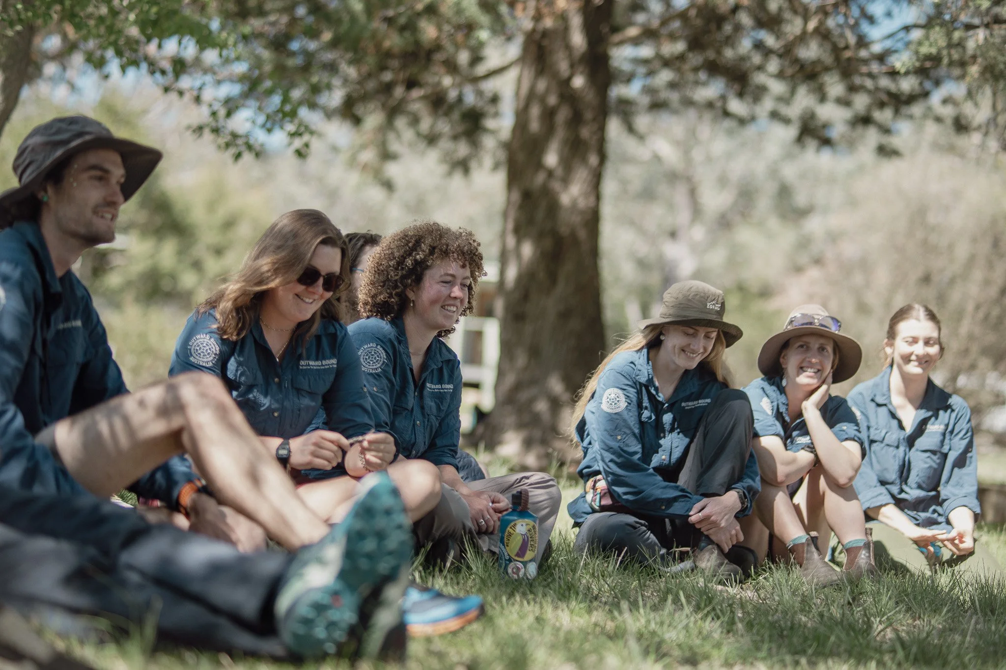 A group of seven women sitting on grass outdoors in front of a large tree, smiling and laughing.