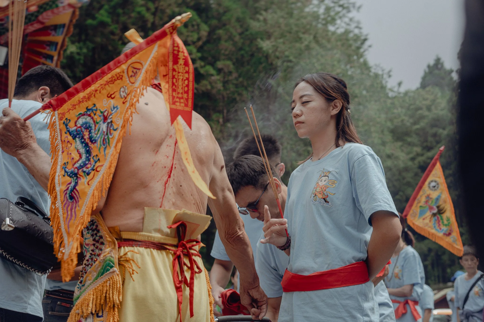 A woman with a red sash around her waist holding incense sticks during a religious or cultural ceremony outdoors, with a shirt embroidered with a mythological figure, surrounded by other participants and colorful flags with intricate designs.