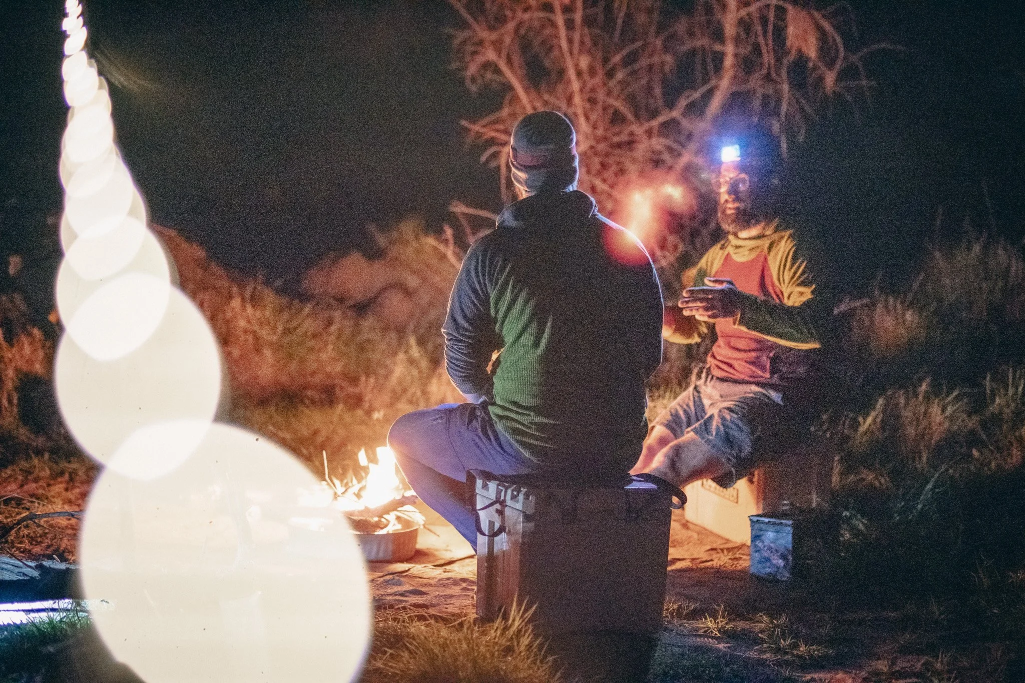Two people sitting around a campfire at night, one wearing a headlamp, with a tree and dry grass in the background.