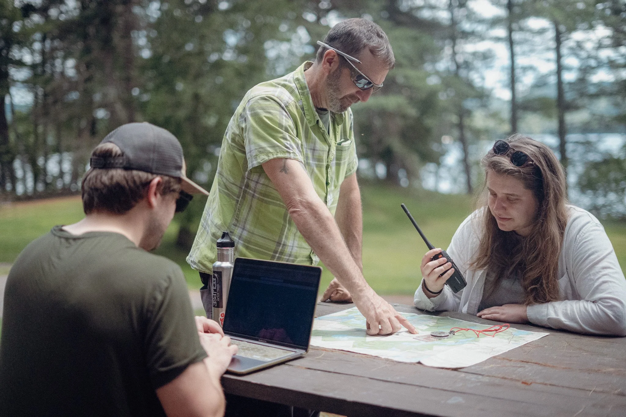 Three people outdoors at a picnic table, looking at a map and using communication devices, with a wooded background and a body of water in the distance.
