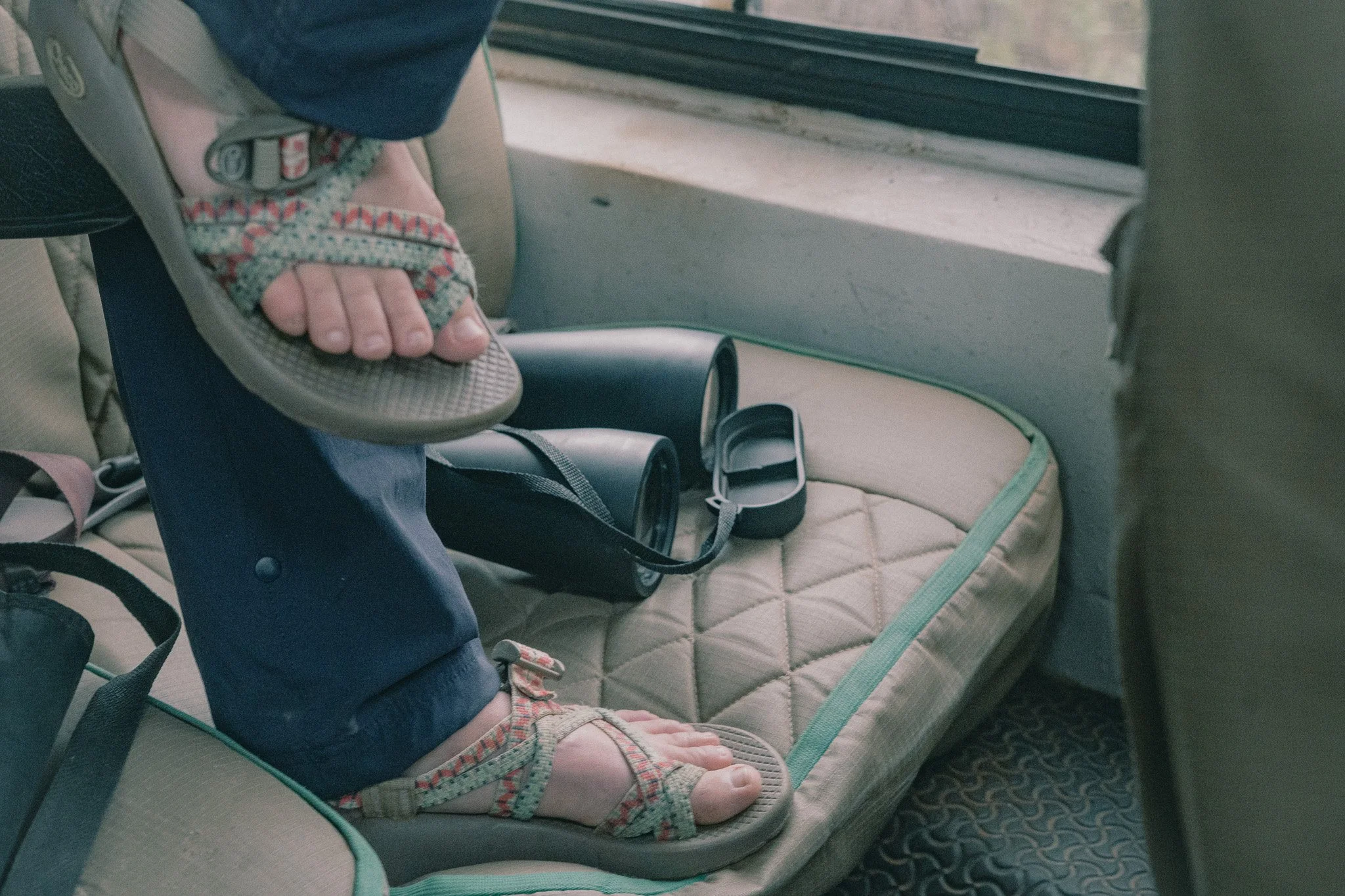 Child’s feet in sandals resting on a cushioned bus seat next to a window, with a bottle holder nearby.
