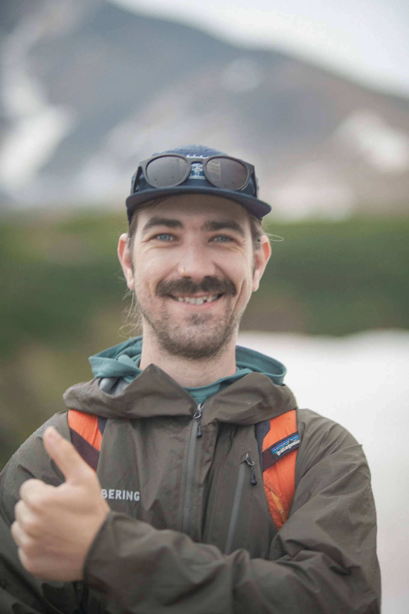 Smiling man wearing outdoor gear, including a jacket, cap with sunglasses, and a backpack, outdoors in a natural setting.