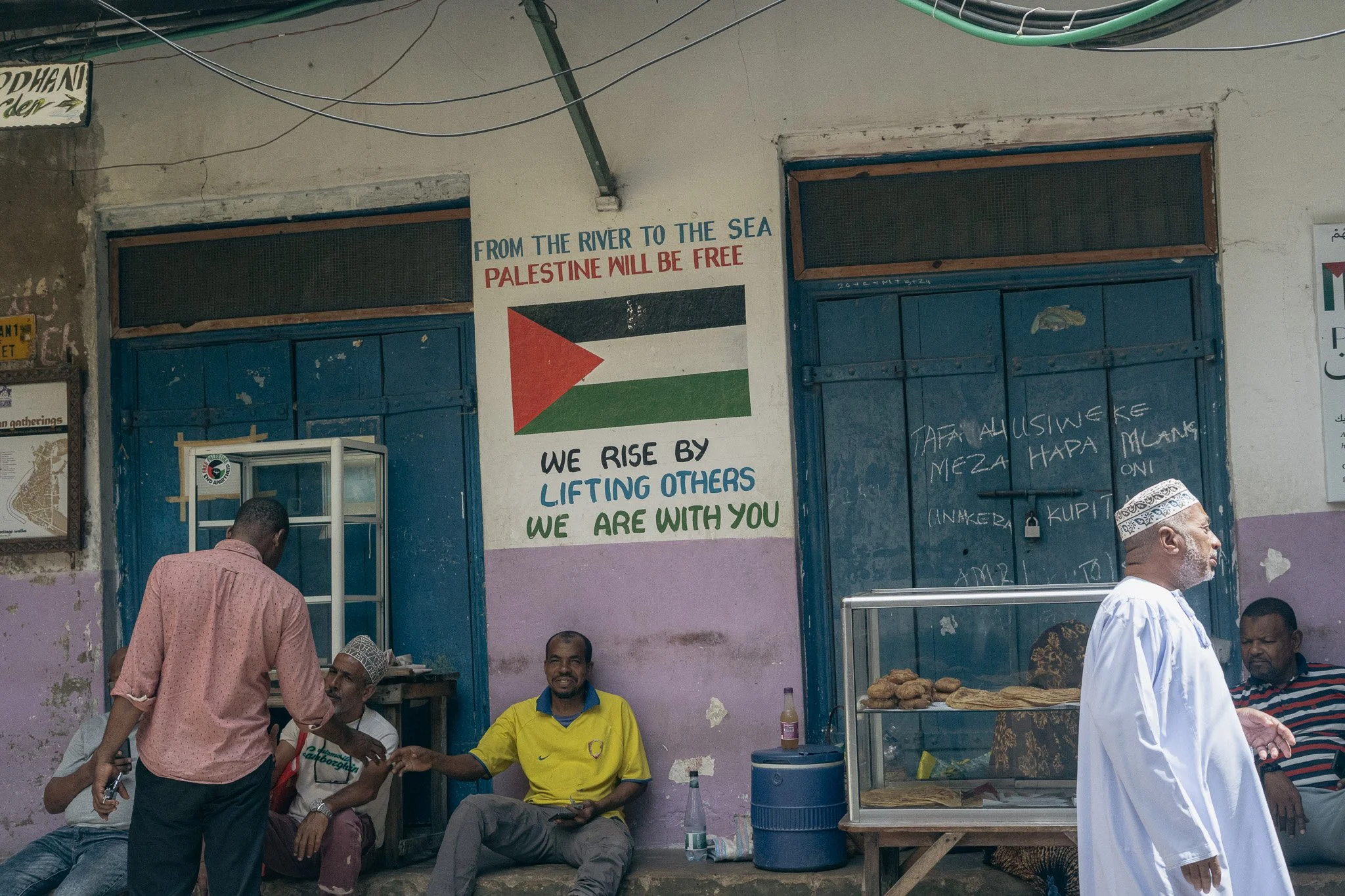 Street scene with four men sitting and standing in front of a building. One man is handing something to another. The building has a painted sign with a Palestinian flag and the message 'From the river to the sea, Palestine will be free. We rise by li
