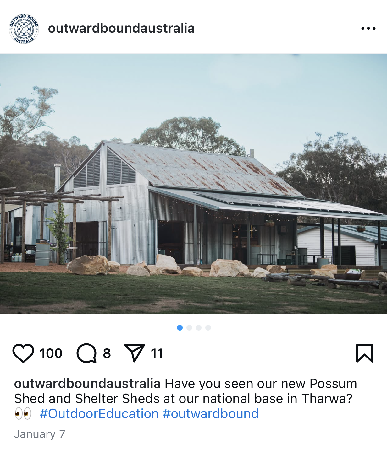Rustic metal shed with a sloped roof and a covered porch area in an outdoor setting, surrounded by rocks and greenery, with a background of trees and a clear sky.