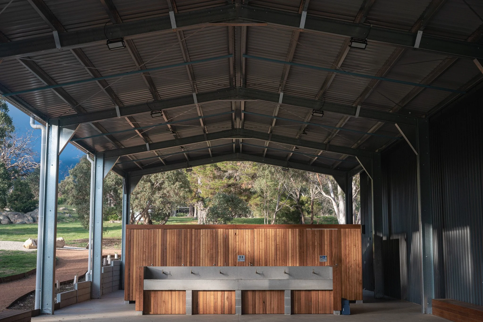 Outdoor park shelter with metal roof, wooden and concrete picnic tables, surrounded by trees and grass.