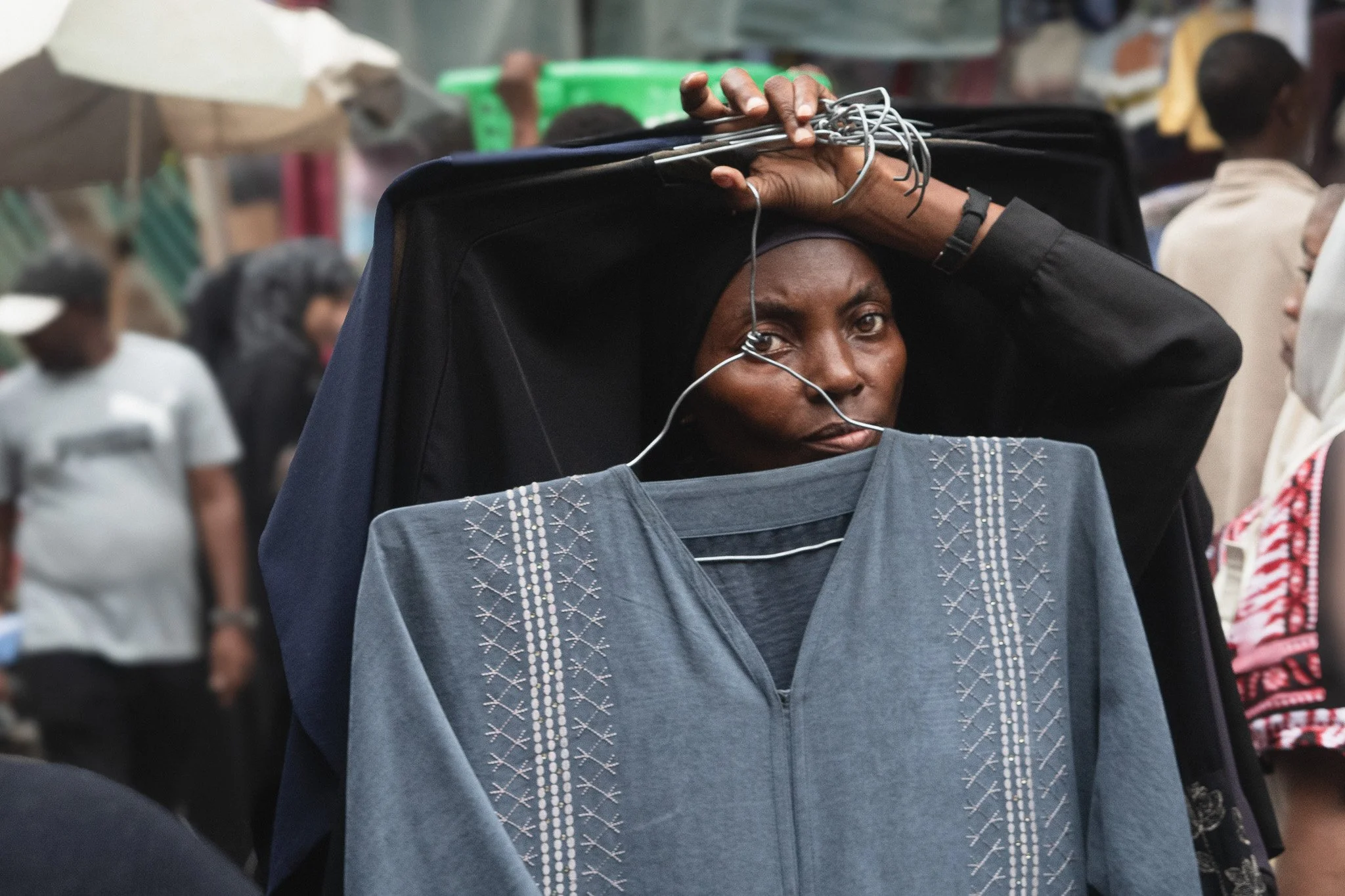 A woman holding a blue garment on a hanger over her neck, with another garment draped over her head, standing in a crowded market.