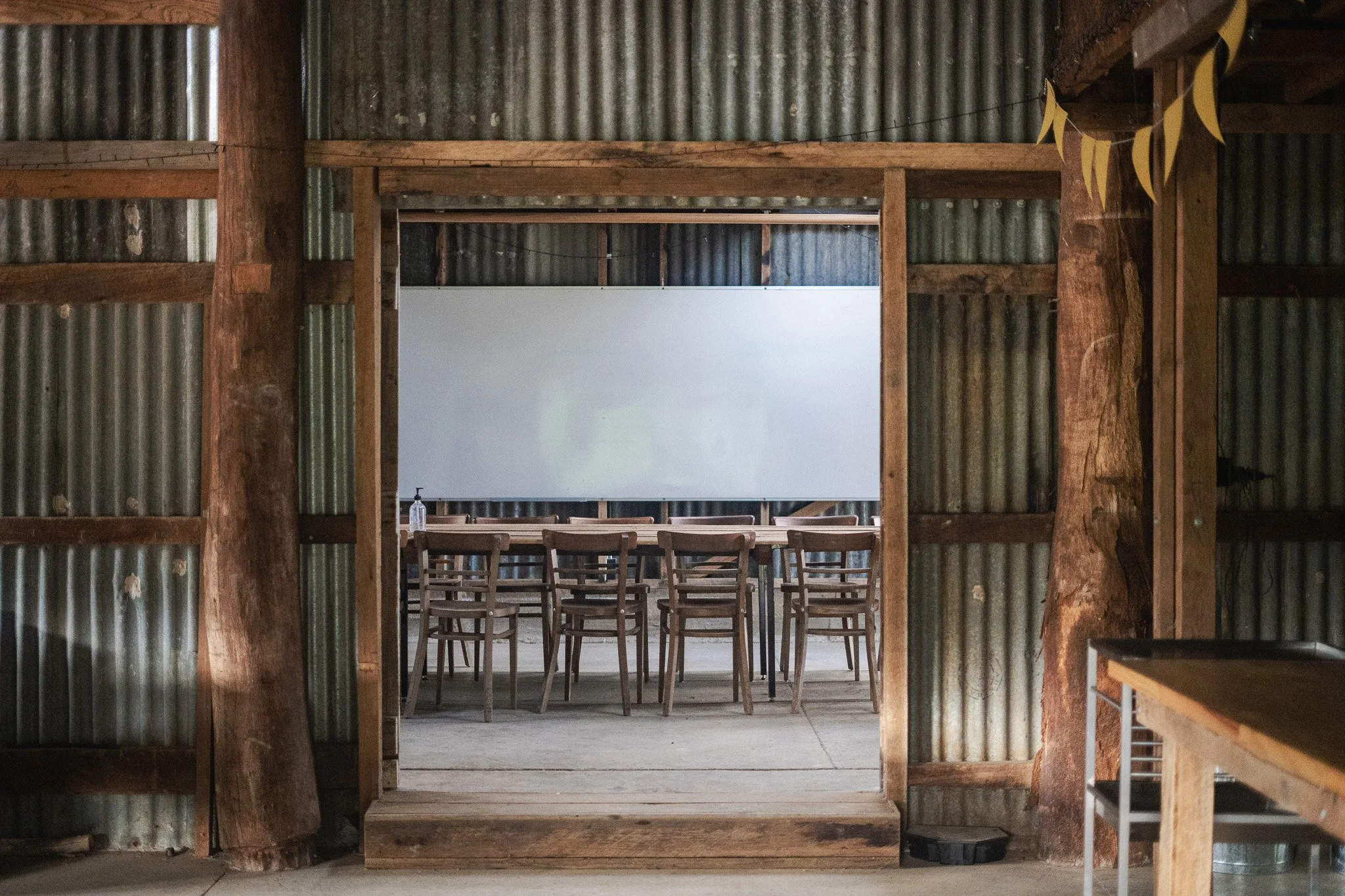View through a rustic barn doorway into a workshop or meeting area with wooden table, chairs, and a whiteboard or screen on the back wall.