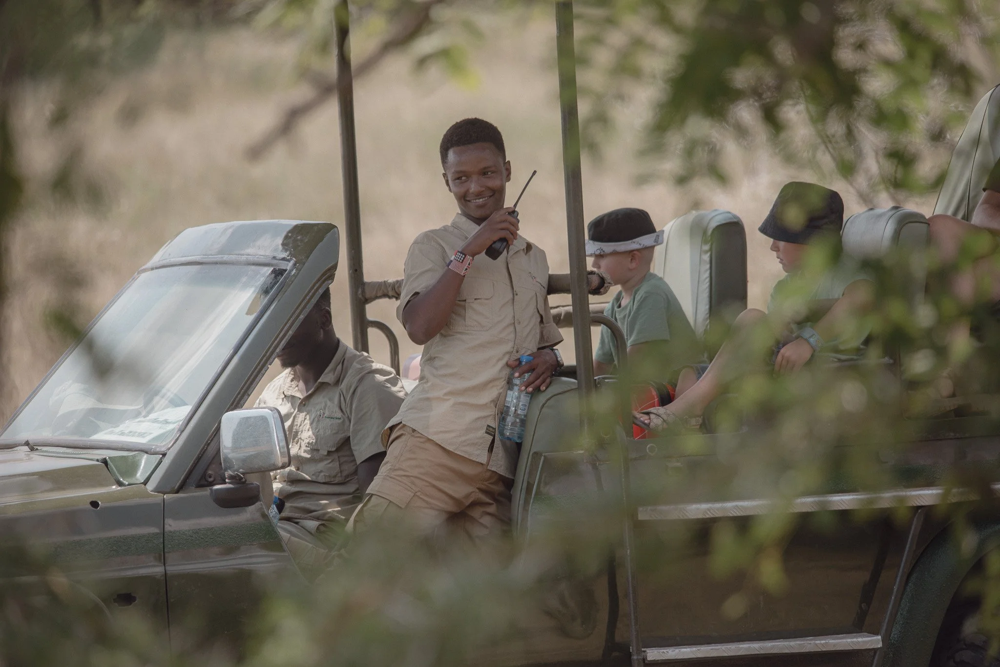 A young male park ranger with a radio in hand smiling while standing in an open safari vehicle with children seated inside, surrounded by trees.